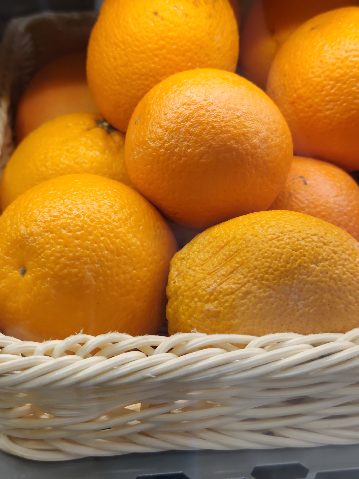 A close-up, eye-level shot captures a woven white basket filled with bright orange tangerines. The tangerines are clustered together, some overlapping, with their bumpy skins illuminated by an unseen light source. The texture of the fruit's peel is a prominent feature. The basket is filled to the brim, with only the top portions of the tangerines visible, creating a sense of abundance. The background is blurred and dark, emphasizing the foreground subject. There are no people or actions depicted in the image. The scene appears to be indoors, possibly in a market or store. The location in Sochi, Russia, is not discernible from the visual information provided.