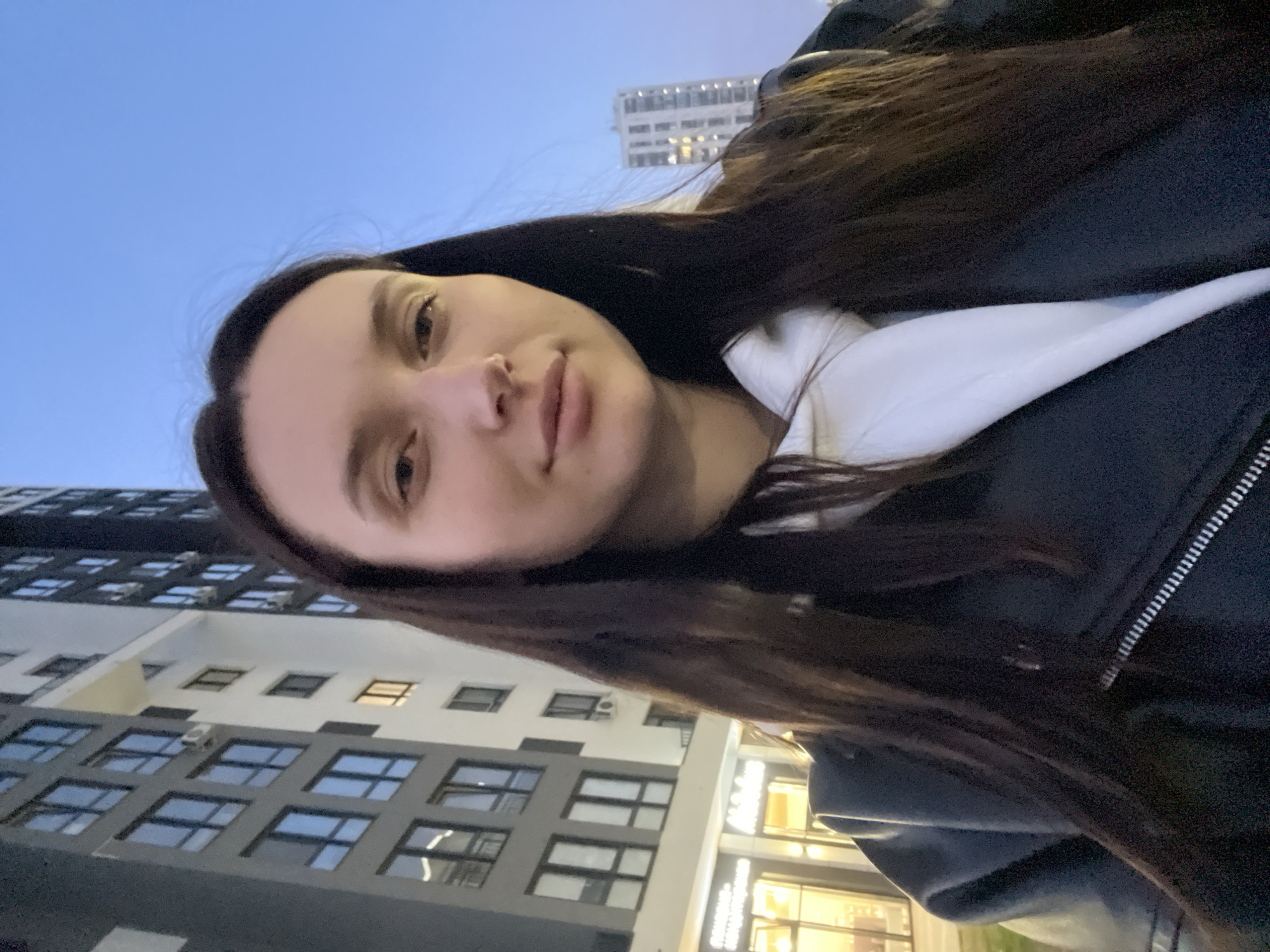 A person with long, dark brown hair is taking a selfie outdoors. They are wearing a white hooded sweatshirt under a dark jacket. The background shows city buildings at dusk or night. The building on the left has multiple windows with a gray and white exterior. In the distance, another taller building is visible. The image appears to be taken in an urban area. Based on the location of the building in the background, the image was likely taken in the city of Tyumen, Russia.