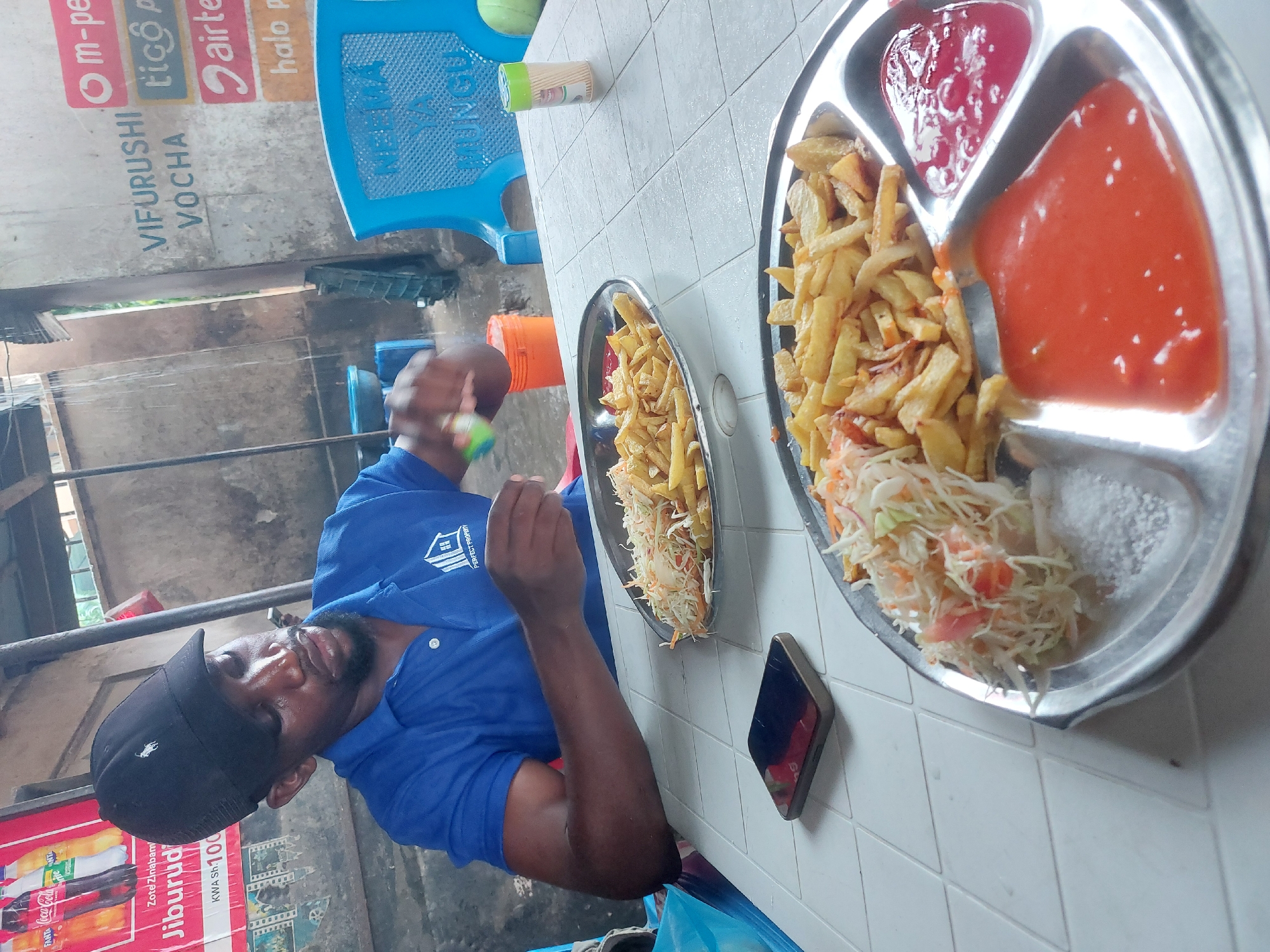 A man is seated at a table in what appears to be an outdoor or semi-outdoor eatery in Dar es-Salaam, Tanzania. He is wearing a blue polo shirt with a company logo "PERFECT PROPERTY" and a black baseball cap. He holds a green object in his right hand, possibly food or a condiment.
Two plates of chips are on the table. The larger plate is sectioned, containing piles of french fries, salad, ketchup, hot sauce, and salt. A mobile phone lies on the table between the man and the plates.
In the background, promotional posters advertise mobile service providers (m-pesa, tico P, airtel, halo p) and a local beverage (Jiburud). There are blue plastic chairs and a white-tiled table. The backdrop suggests a simple, open-air dining setting, typical of local eateries in Dar es-Salaam.