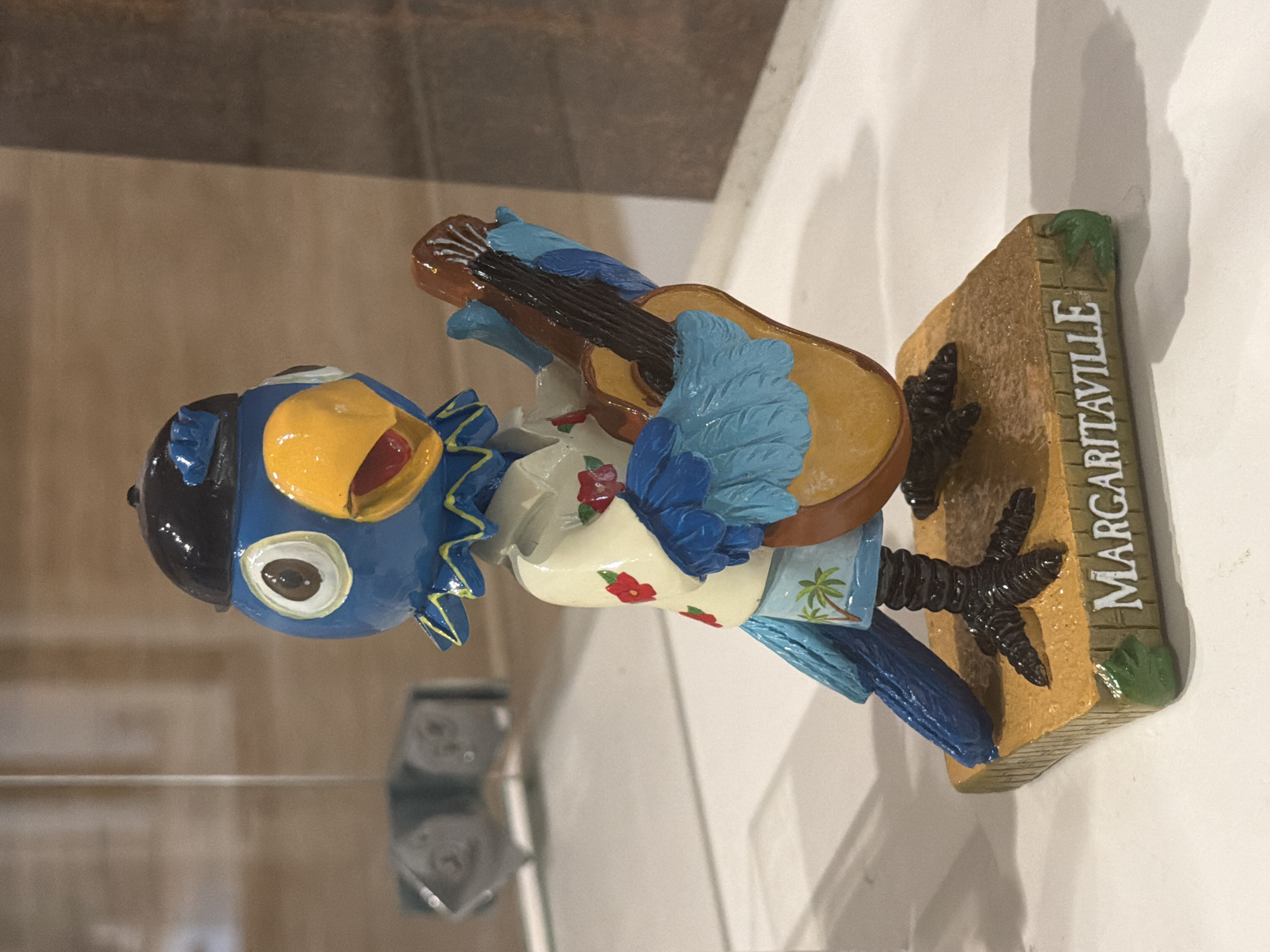A close-up, eye-level shot captures a decorative figurine of a blue bird wearing a black hat and a Hawaiian-style shirt, playing a brown guitar. The bird stands on a square, tan-colored base that reads "MARGARITAVILLE" in white, stylized lettering. The base also features green painted leaves on two of its sides. The background is blurred, showing a vertical white pole to the left and a textured, brown surface to the right. The figurine is lit from the front, casting a shadow to the right of the base.
