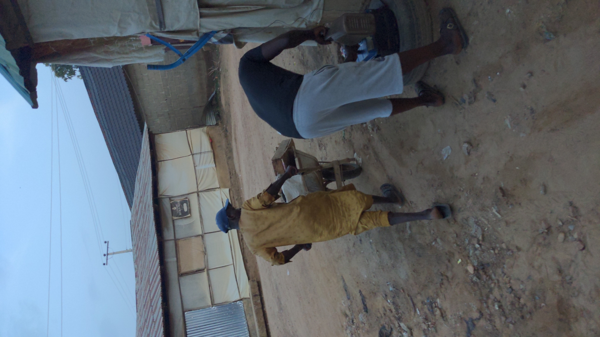 Two people stand outside a building constructed from a patchwork of materials, including corrugated metal, cloth, and wood. The man on the left wears a blue cap and a long, mustard-colored robe, standing on the sandy ground. To the right, a person in a black shirt and light-colored shorts appears to be interacting with a piece of machinery. The setting appears informal and somewhat improvised, with a mix of temporary and more permanent structures. A power pole with electrical lines stands near the building.
