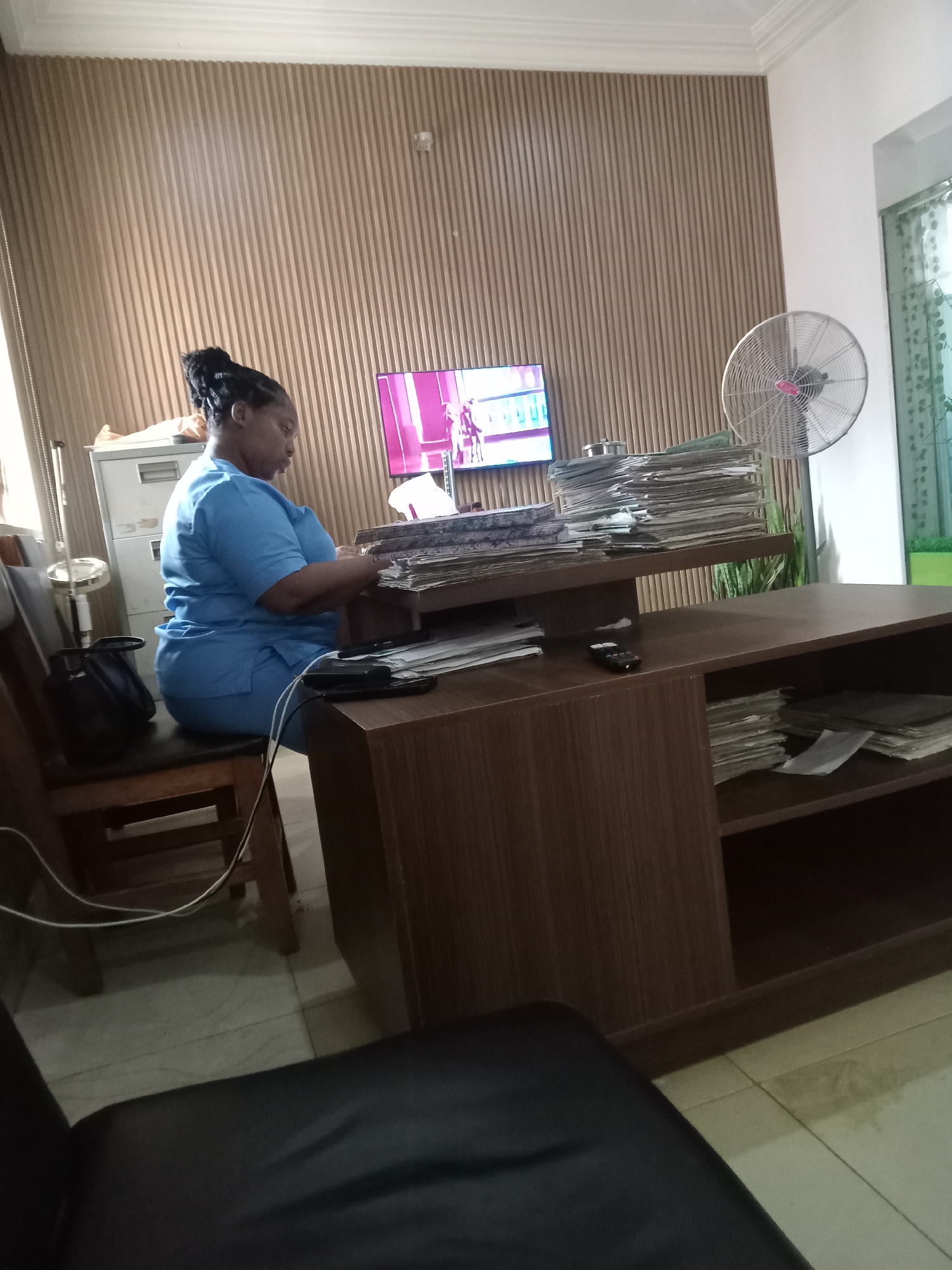 The image depicts a woman in a light blue scrub suit sitting at a brown wooden desk in an indoor setting. She has dark hair pulled back in a bun and appears to be working on a large stack of papers. The desk has a raised platform, also covered with papers, and shelves below containing more papers. A black bag hangs from the back of the wooden chair. There are two black objects that appear to be phones on the desk. Behind her, a filing cabinet is visible to the left of a flat-screen television mounted on a wooden slat wall. To the right of the television is a standing electric fan. A green potted plant is positioned near a window. The location is presumed to be Gwarinpa, Nigeria.