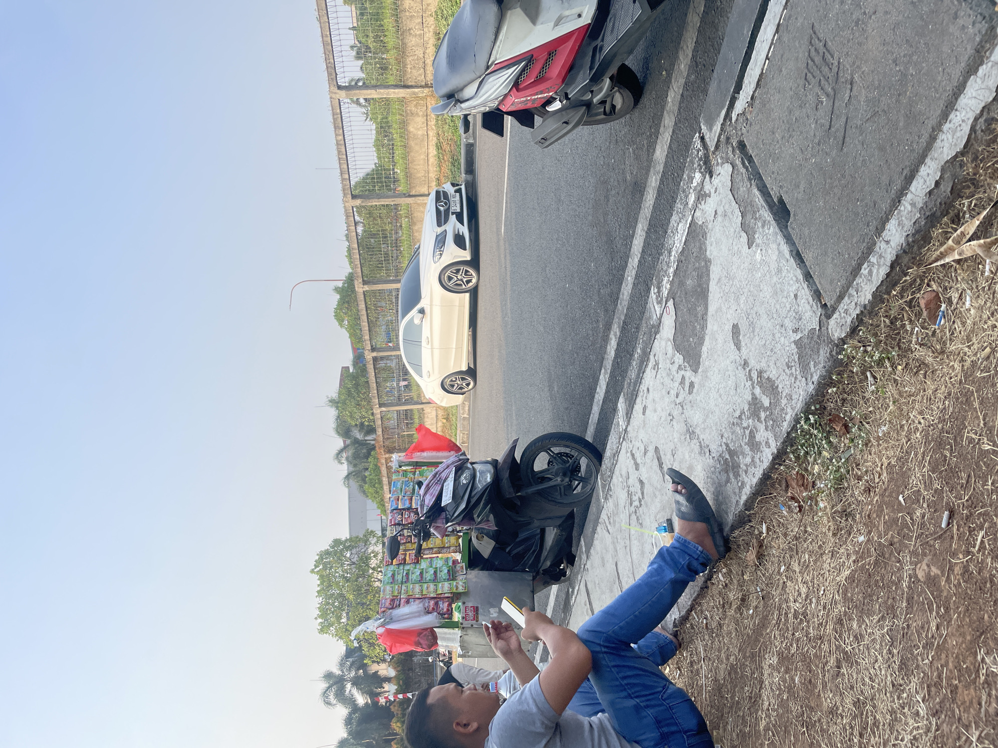 A man sits on the side of a road in Danauindah, Indonesia, holding a smartphone and smoking. He is wearing a gray t-shirt and blue jeans. Beside him is a motorbike with a cart attached, displaying various goods for sale, likely snacks or cigarettes. The cart is adorned with red flags. In the background, a white Mercedes-Benz sedan is parked on the road, and another motorbike is visible. The scene appears to be in an urban or suburban area with a sidewalk, a road, and a concrete barrier separating the road from a grassy area. The sky is clear and the lighting suggests it is daytime.