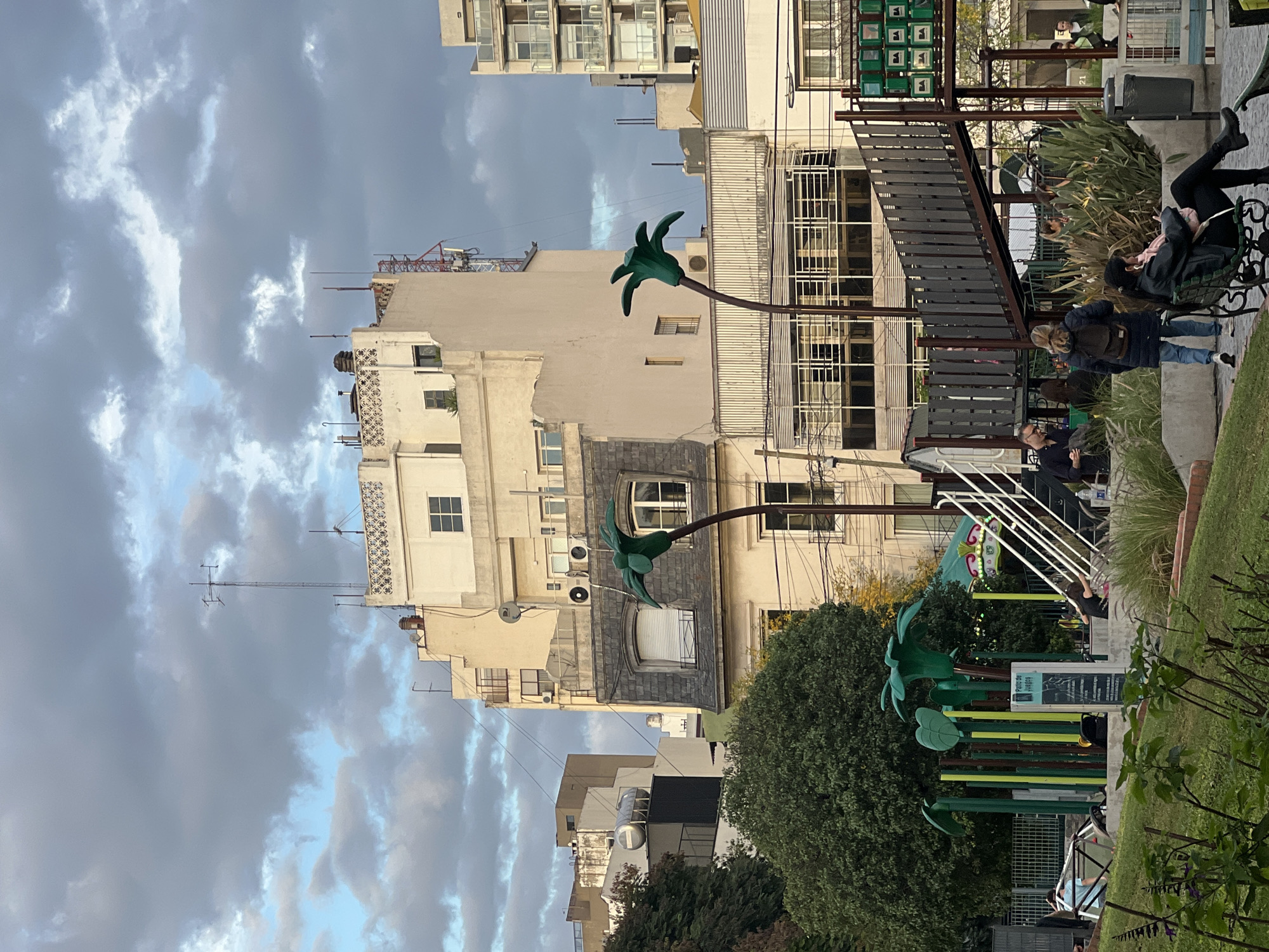The image depicts a cityscape in Buenos Aires, Argentina. The sky is overcast with gray clouds. In the foreground, there are green, artificial palm tree sculptures made of metal. People are sitting on benches in what appears to be a park. Buildings of varying heights and architectural styles form the backdrop. The buildings are mostly light-colored and include apartment blocks and structures with visible antennas and satellite dishes. Some buildings have ornate facades. In the right side of the image there is what appears to be a merry-go-round and people sitting. The photo is taken in a somewhat close range, capturing a mix of urban architecture and recreational space.