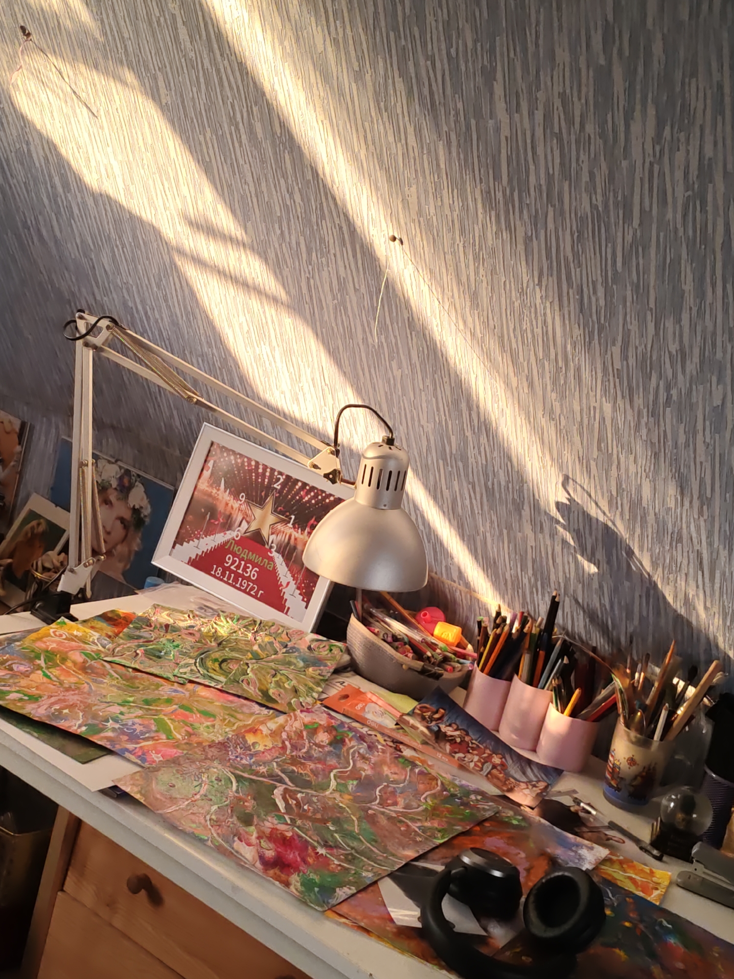 The image depicts an art desk bathed in dappled sunlight, likely in a residential setting in Rostov-on-Don, Russia. The wall behind the desk is covered in textured wallpaper, illuminated by diagonal shafts of light cast by an unseen window.
On the desk, a white architect's lamp with exposed springs is positioned to the left. In front of the lamp is a framed picture displaying a red carpet, a star, and text that appears to be a name and a date in Russian, "Людмила 92136 18.11.1972 г". To the right of the framed picture, a variety of art supplies are arranged. A grey fabric bin is filled with colored pencils and markers. Three pink cylindrical containers hold more colored pencils and pens. A clear glass jar contains an assortment of paintbrushes.
Scattered across the desk are several pieces of abstract artwork, characterized by vibrant, swirling colors and organic shapes, suggestive of fluid art techniques. In the foreground, a pair of black headphones rests on top of some of the artwork. Further to the right, a small glass sphere, possibly a snow globe, sits next to a silver stapler. The desk itself appears to be made of wood, with visible drawers and a pull knob. The overall impression is that of a creative workspace in use, bathed in the warm light of late afternoon or early morning.
