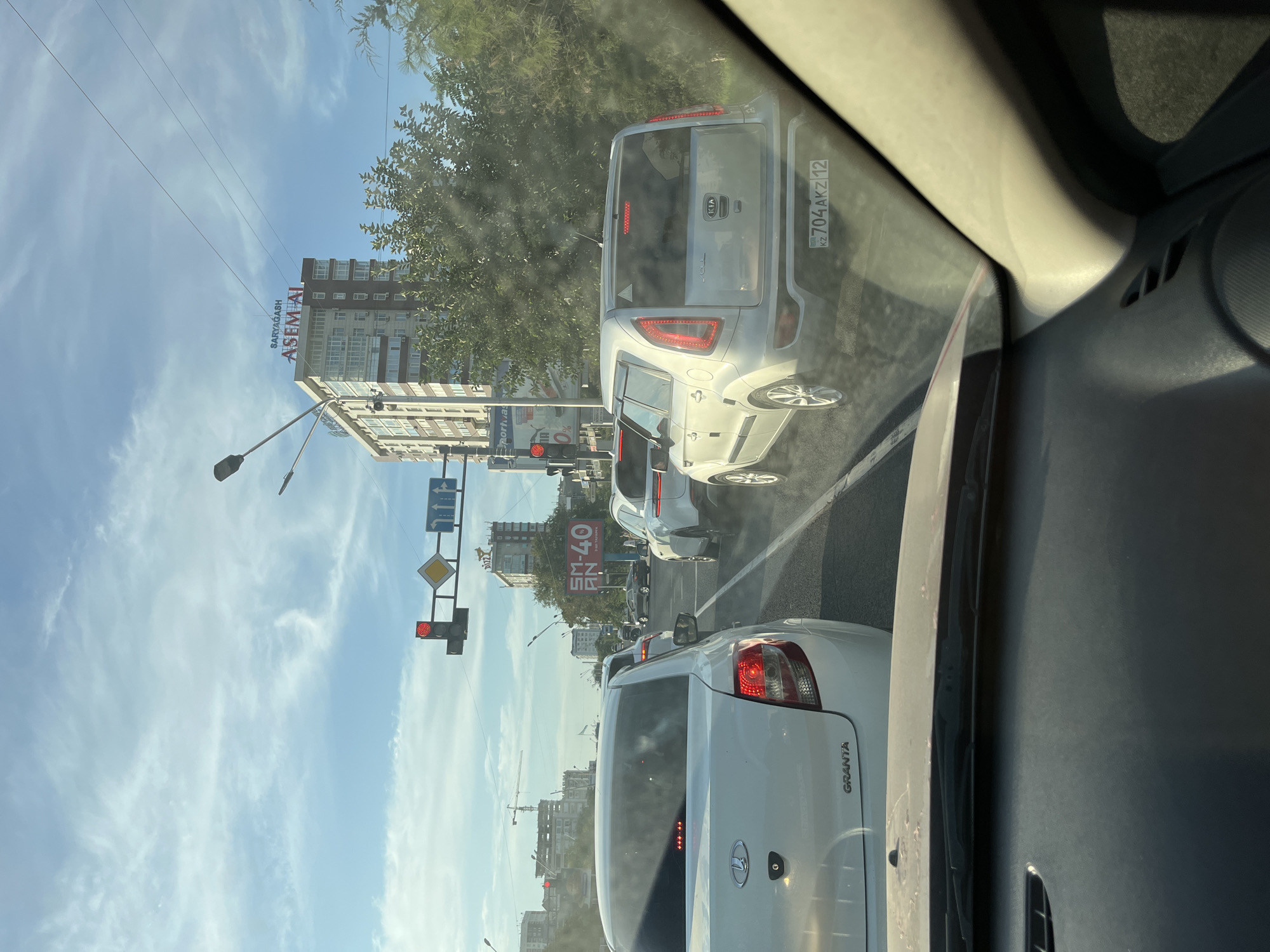The image depicts a traffic scene in Aktau, Kazakhstan, likely on a sunny day with scattered clouds. The foreground shows the interior of a car, with the dashboard and windshield visible, suggesting the photo was taken from the driver's perspective. The windshield is smudged, indicating it is not perfectly clean.
Ahead, multiple cars are lined up, suggesting a traffic jam or waiting at an intersection. The cars include a white Lada Granta in the immediate foreground, a silver Kia Soul behind it, and other vehicles further up the road. The license plate of the Kia Soul is clearly visible, reading "KZ 704AKZ 12".
Traffic lights are visible, one of which is red, indicating vehicles should be stopped. Road signs are also present, including a diamond-shaped sign and directional arrows. In the background, modern buildings and trees line the street. One prominent building has signage that reads "SARYAGASH ASEM AI" in large letters. Another billboard displays "SM-40". Construction cranes are also visible in the distance, indicating ongoing development. The overall scene suggests a typical urban traffic situation in Kazakhstan.