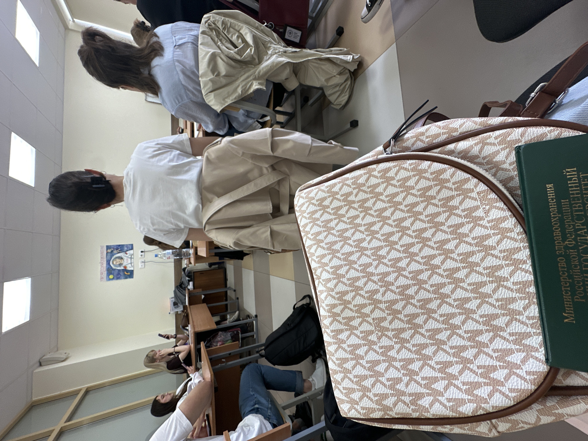 This image captures an indoor scene within what appears to be a classroom or lecture hall, likely in Kazan, Russia. The perspective is from a seated position among students, looking forward towards the front of the room.
In the immediate foreground, a beige and white patterned bag, featuring a repeating "MK" logo, rests on a desk or chair. Partially obscuring the bag is a green book with Cyrillic text. The legible portions read "Министерство здравоохранения Российской Федерации" (Ministry of Health of the Russian Federation) and refer to a "государственный университет" (State University), indicating a medical or health sciences educational institution.
Several young people, presumably students, are seated at wooden desks with metal frames. Most are seen from behind. The person closest to the camera wears a white t-shirt and has dark hair, while the student in front wears a light blue striped shirt and has brown hair tied back. Further down the rows, additional students are visible, some appearing to be engaged with their mobile phones. A black backpack is tucked under one of the desks.
The walls are light-colored, and rectangular white fixtures, possibly lighting panels or projector screens, are visible. A poster on the far wall features an astronaut and the name "Ю. А. Гагарин" (Y. A. Gagarin), a notable Russian figure, reinforcing the location context.
The scene depicts students attending a class or lecture. The time of day is not discernible, but the indoor setting suggests active academic hours. No distinct emotions are evident from the visible subjects.