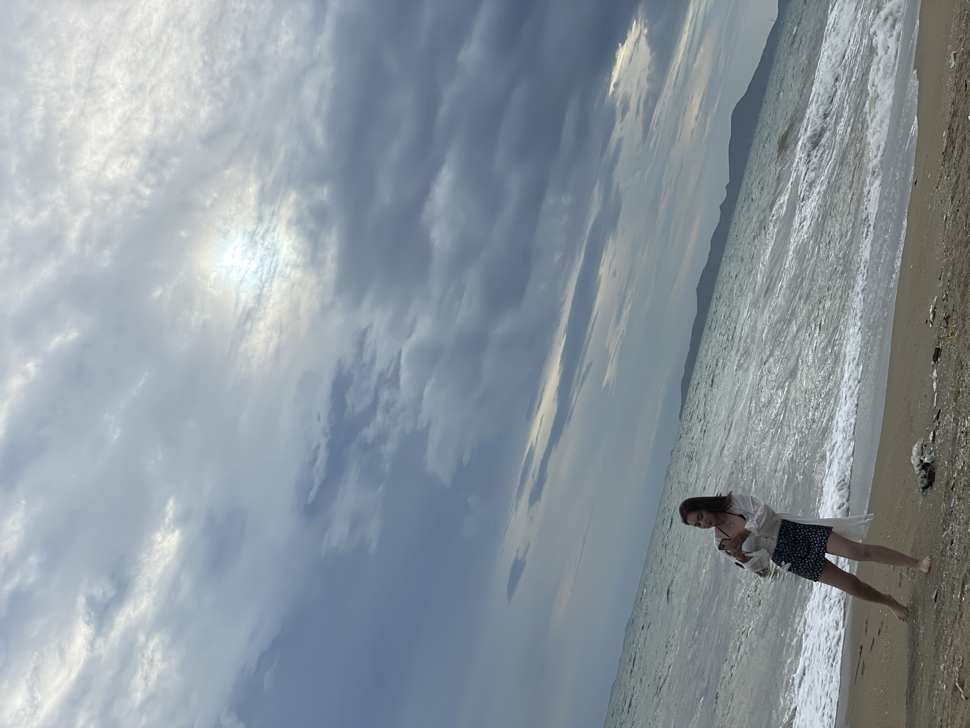 A young woman stands on a sandy beach in Pattaya City, Thailand, with the ocean and a cloudy sky in the background. She is wearing a white sheer cover-up over a black and white patterned skirt and a dark bikini top. She is holding a smartphone in her hands and appears to be taking a photo or video. The waves are crashing on the shore, and the sky is filled with dramatic, dark clouds with a hint of sunlight breaking through. In the distance, the silhouette of mountains can be seen. There is some debris scattered on the sand near the woman.