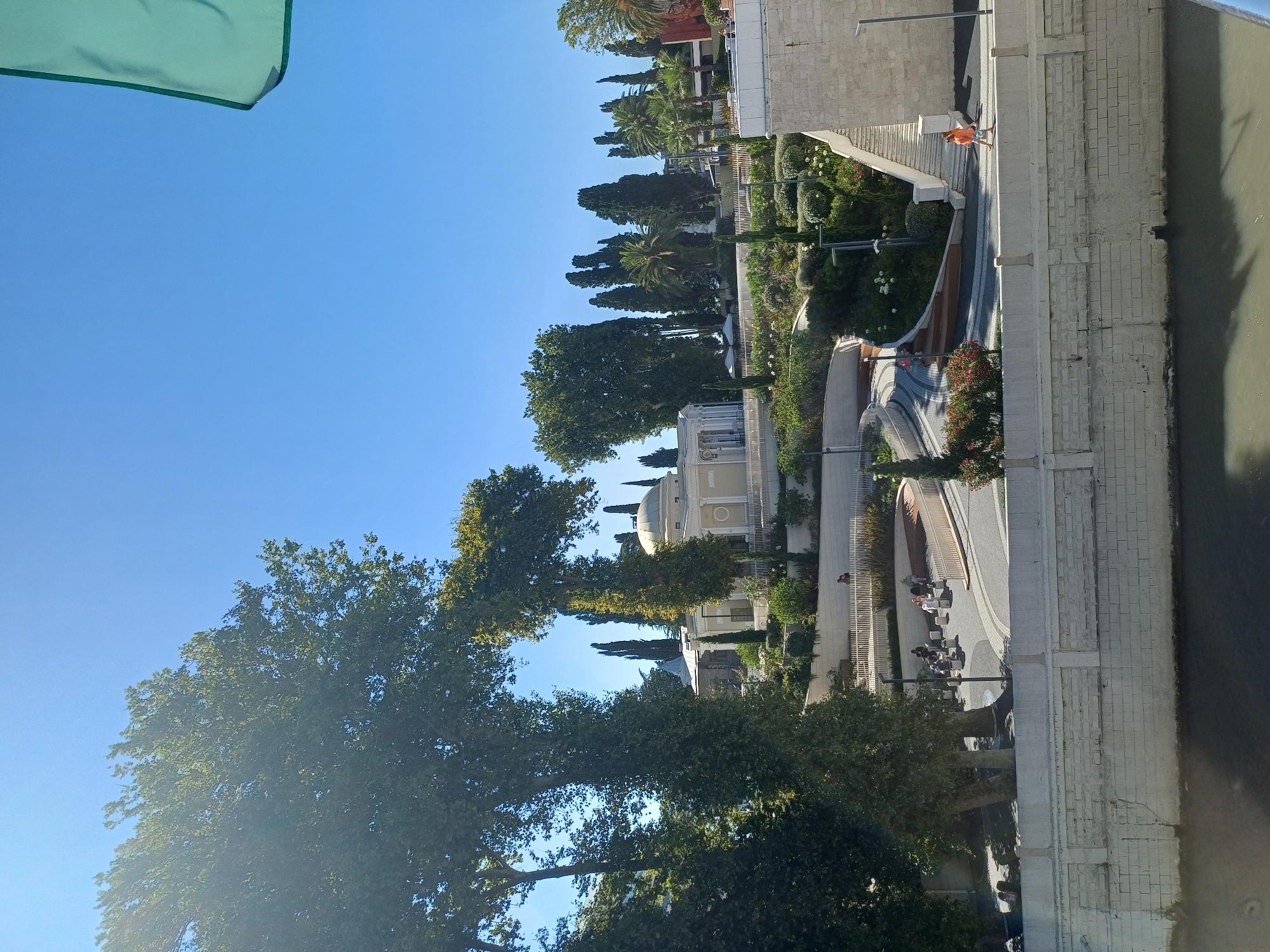 The image shows a scenic view of the Arboretum in Sochi, Russia. A large building with a dome and classical architectural elements is visible in the background, surrounded by lush greenery and tall trees. A winding pathway leads up to the building, with people casually strolling and sitting on benches. The foreground features a stone wall overlooking a body of water, likely a river or the sea, with reflections of the trees and sky. The overall scene conveys a peaceful and idyllic atmosphere, typical of a well-maintained park or garden.