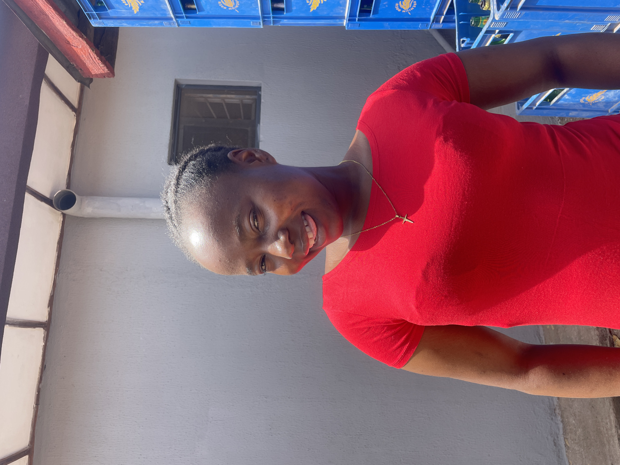 A medium, close-up, angled shot depicts a Black woman in a red dress smiling towards the camera. She has short braided hair, and wears a gold necklace with a cross pendant. Behind her is a plain grey building with a small window and a drainpipe. Blue plastic crates, possibly containing bottled beverages, are stacked near the top right corner of the frame. The location is potentially Jos, Nigeria, based on the prompt.