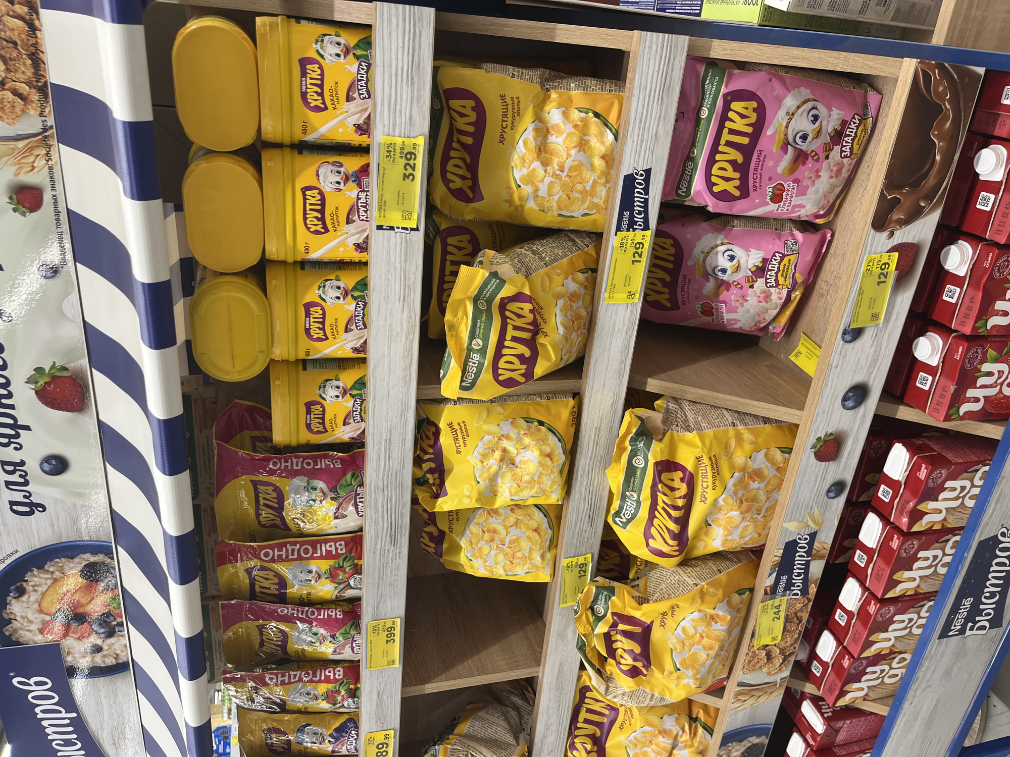 The image shows a retail display of breakfast cereal products in a supermarket. The products are arranged on wooden shelves, with various brightly colored packaging. Several brands are visible, including "ХРУТКА" (Khrutka) and "Nestle Быстров" (Nestle Bystrov). The products include what appear to be corn flakes, and some are flavored with strawberry and vanilla, or cocoa. There are price tags visible, with discounts indicated. In the background, there are more shelves and products, suggesting a typical supermarket environment. The image was taken in Volzhsky, Russia.