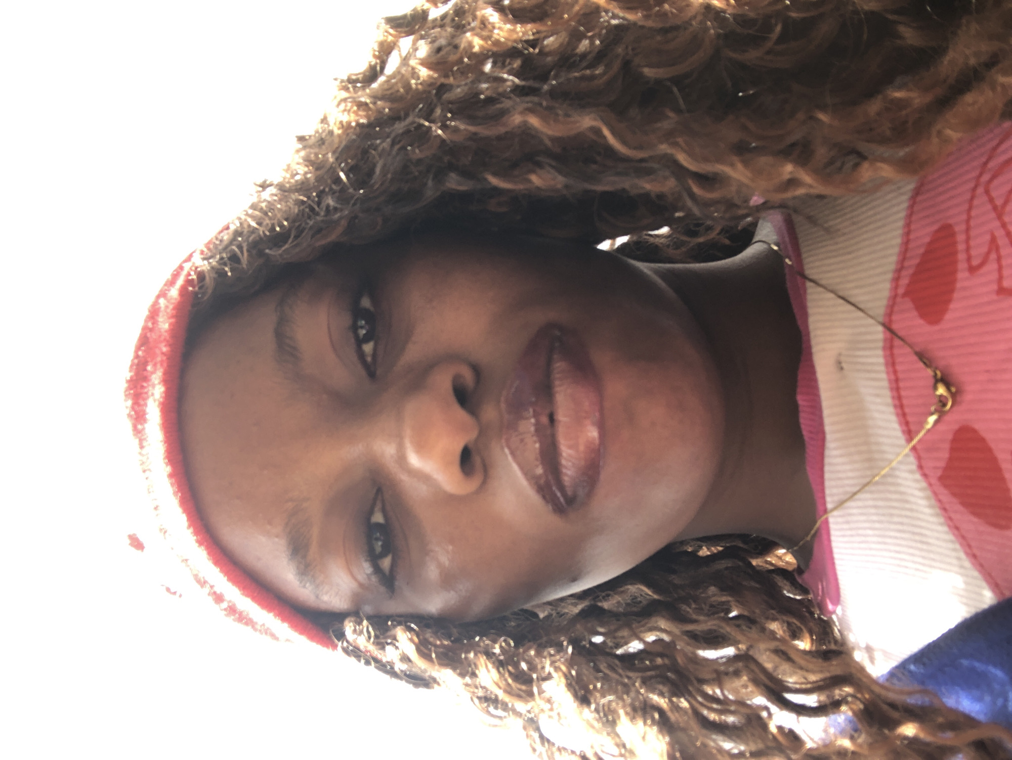 A close-up, slightly angled portrait of a person with dark skin, full lips with glossy lipstick, and dark eyes. They are wearing a red headband and have curly brown hair. A thin gold chain necklace is visible. The person is wearing a top that is patterned with pink and white. The background is predominantly bright and overexposed. The location is not discernible as the background is overexposed and indistinct, and no identifying markers are present. It is therefore impossible to confirm the location as Pindiga, Nigeria.
