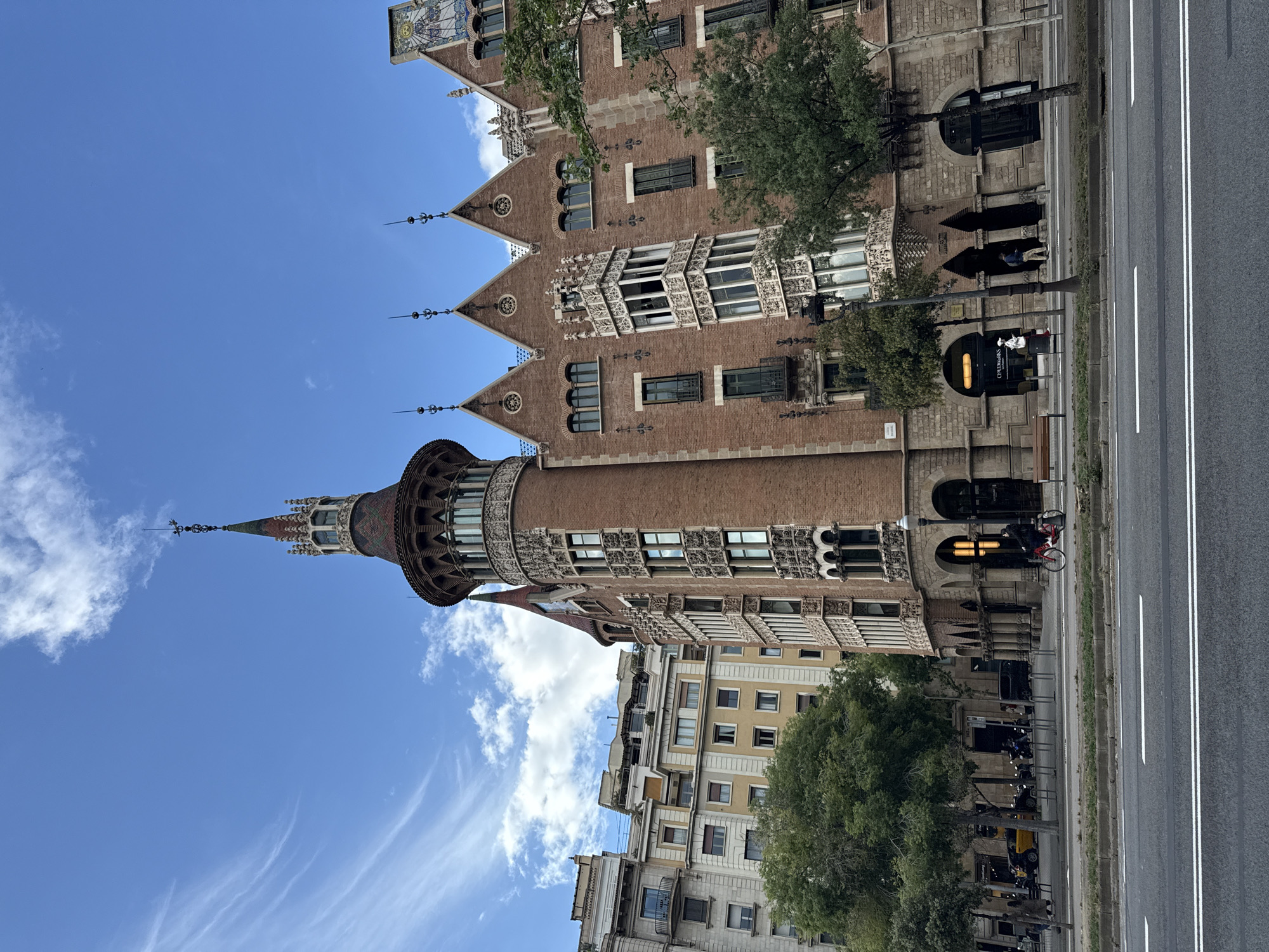 The image shows Casa de les Punxes, a Modernista building in Barcelona, Spain. The building is made of red brick and has a distinct turret with a pointed roof. The sky is blue with some clouds. A traffic light and street is in front of the building with a cyclist. Other buildings are visible in the background.