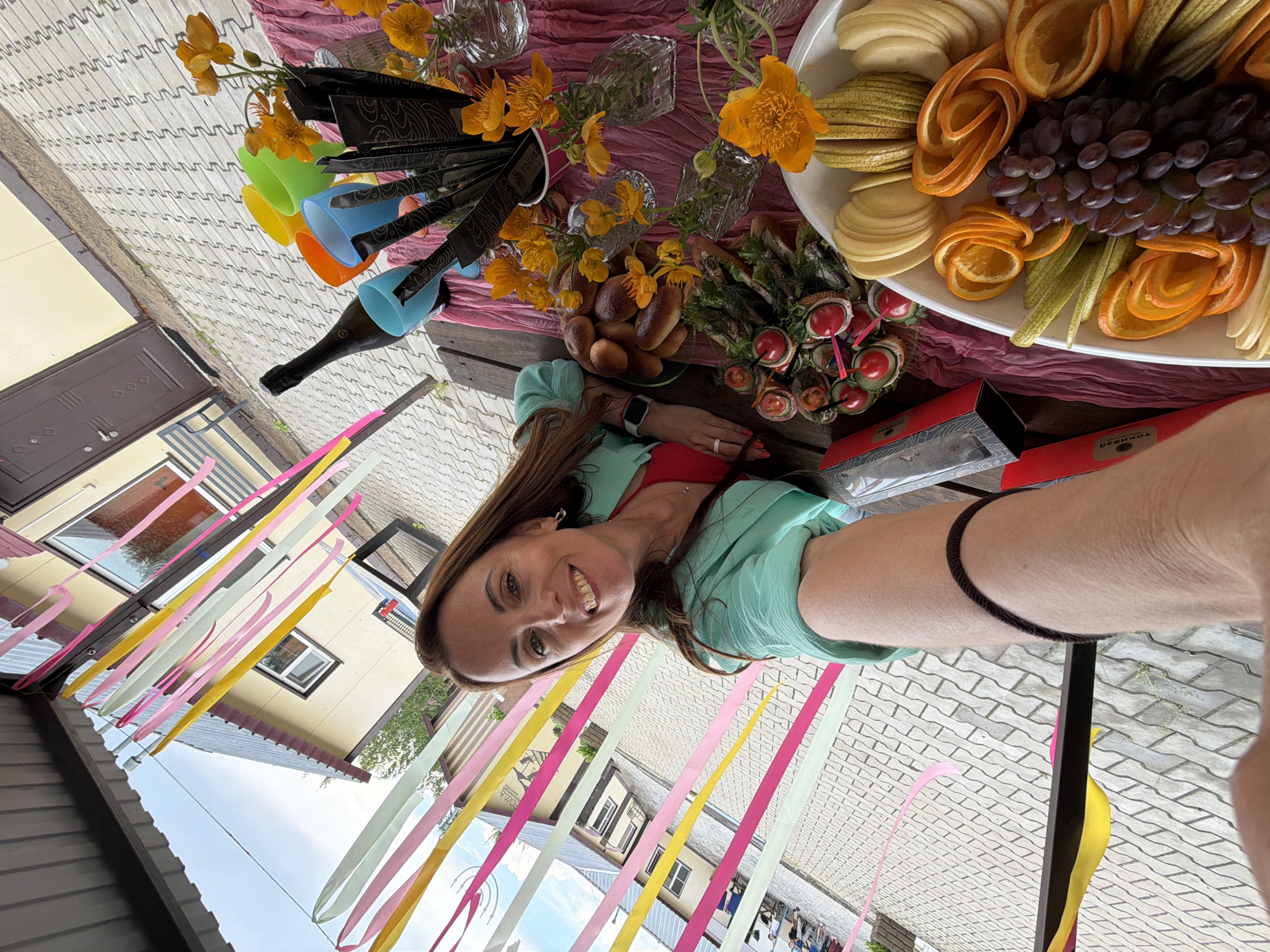 An outdoor selfie captured by a woman with long brown hair, smiling at the camera. She is wearing a teal jacket over a red top. The scene appears to be a celebration or gathering, with a table laden with food and drinks. Visible items include a bottle of champagne, colorful plastic cups, a bouquet of orange flowers, a platter of assorted fruits (grapes, oranges, pears, apples), and small pastries or buns. Decorative streamers in pink, yellow, and light green hang overhead. In the background, a building with a brown door and windows can be seen, and the ground is paved with interlocking gray bricks. The overall setting suggests a bright, daytime event in a residential or public outdoor space. The location is Birobidzhan, Russia.