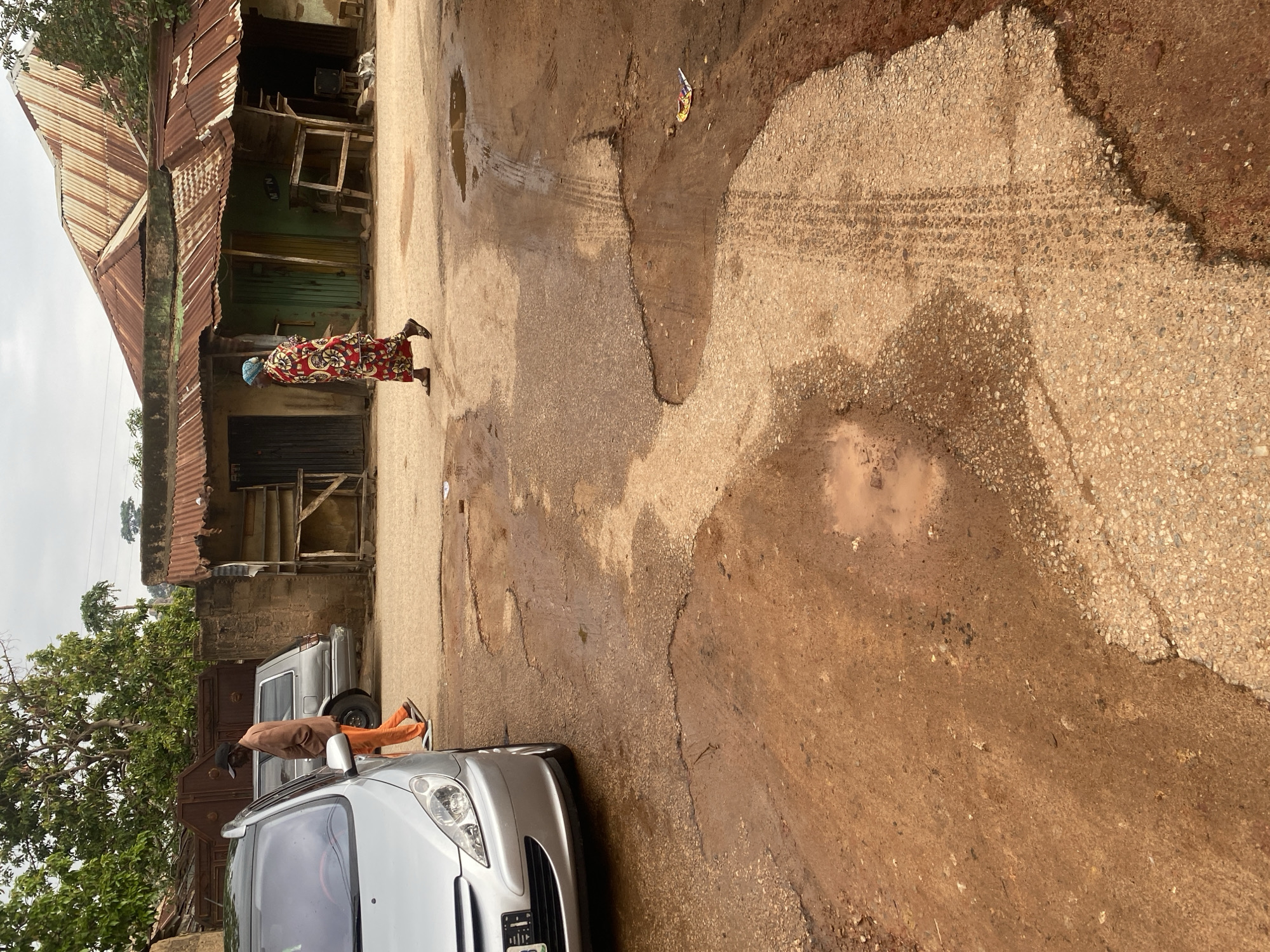 The image captures a street scene in Jos, Nigeria, characterized by a dilapidated environment. In the foreground, a silver hatchback car is parked on a dirt and asphalt road riddled with potholes and puddles of water, indicating poor road conditions. To the left, a man in an orange tracksuit and a black baseball cap walks away from the camera, his attention directed elsewhere. Further down the street, a woman in a vibrant red and yellow patterned dress, with a blue headscarf, walks towards the right. Behind her, a low-rise building with a corrugated tin roof, showing signs of rust and decay, stands as a backdrop. The building appears to house small shops or residences, with some doorways and wooden structures visible. The overall impression is one of an underdeveloped urban area with evident signs of wear and tear.