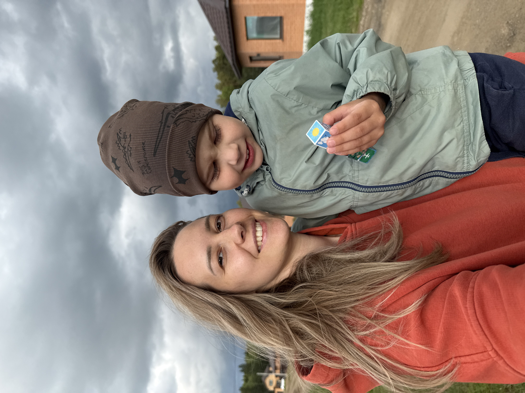 A selfie depicts a woman and a young boy. The woman is positioned at the bottom of the frame wearing an orange hooded sweatshirt and has long blonde hair. The boy is situated above her, wearing a brown hat and a light green jacket. He is holding a cube toy. The background includes a grey, cloudy sky and a small brick building with a window. The photo was taken in Chaikovsky, Russia.