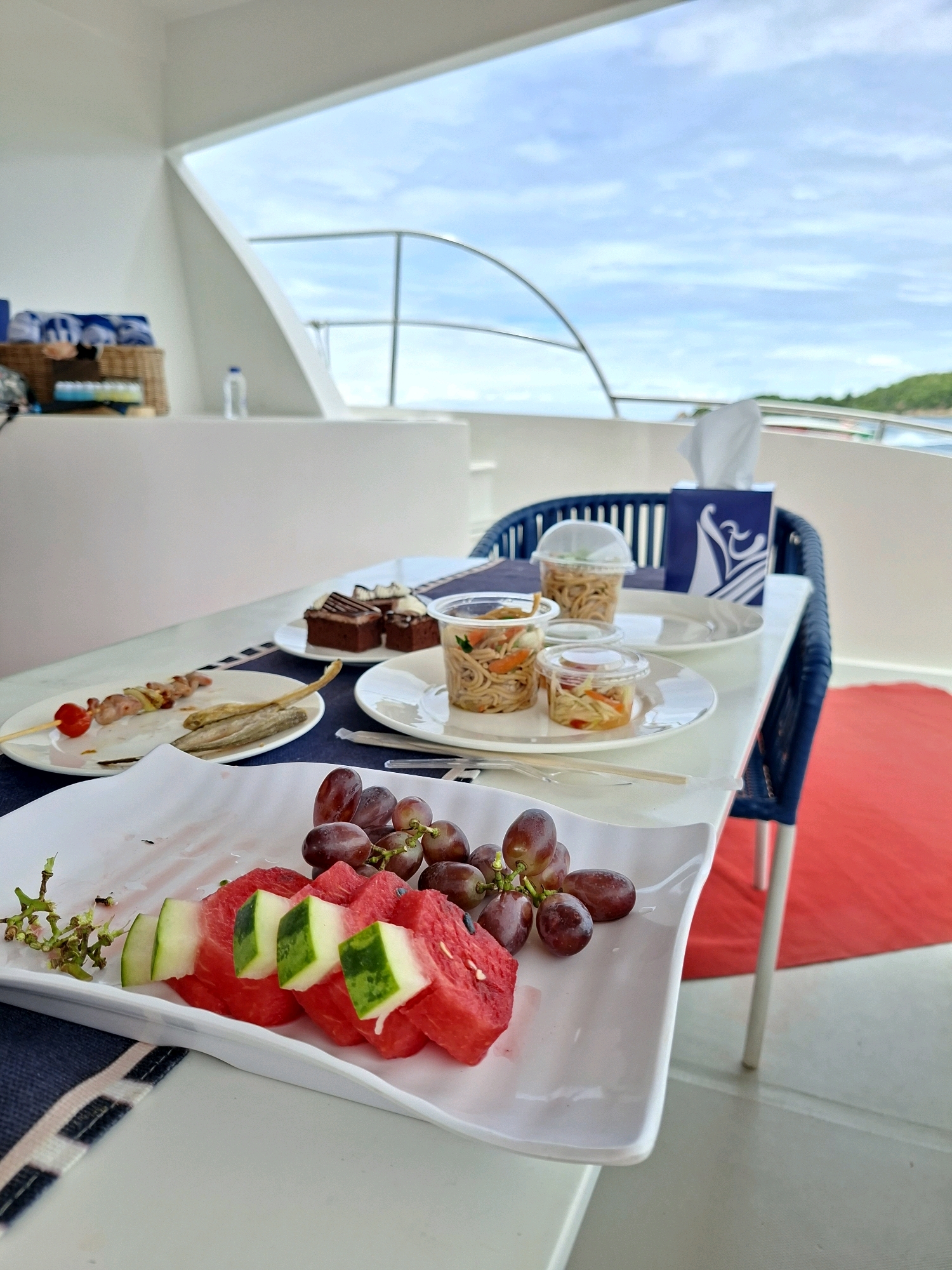 A table set for a meal is positioned on the deck of a yacht, with the ocean and a distant, green, mountainous coastline visible under a cloudy sky. The table holds several plates of food, including sliced watermelon and grapes, small chocolate cakes, and individual servings of noodles in clear plastic containers. A blue napkin dispenser with a white logo sits on the table. A red rug is visible on the deck to the right of the table, and a woven basket containing rolled-up towels can be seen in the background. A bottle of water is also present on a white surface behind the table. The scene is devoid of any people.
