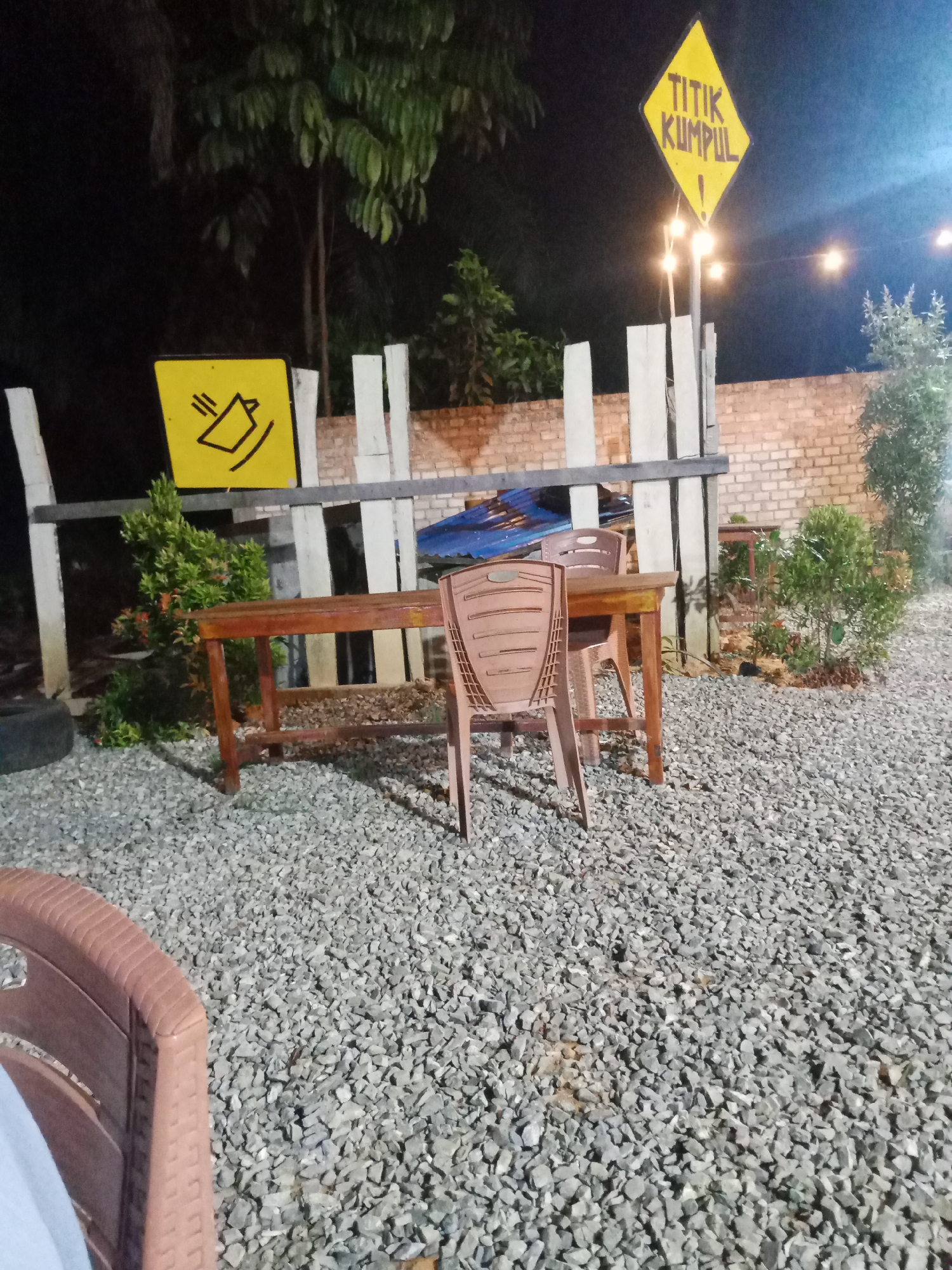 The image depicts an outdoor seating area at night in Mekar Jaya, Indonesia. The ground is covered with small gray gravel. In the foreground, part of a brown plastic chair is visible on the left. Behind it, a wooden table with two brown plastic chairs is set up. A yellow diamond-shaped sign with black text reading "TITIK KUMPUL" and an exclamation mark is mounted on a pole to the right of the table, illuminated by string lights that hang above. To the left of the table, another yellow sign with a black graphic resembling a falling object is displayed. A brick wall forms the background, with a wooden fence and some foliage in front of it. Trees are visible behind the fence. The overall ambiance suggests a casual outdoor dining or gathering spot.