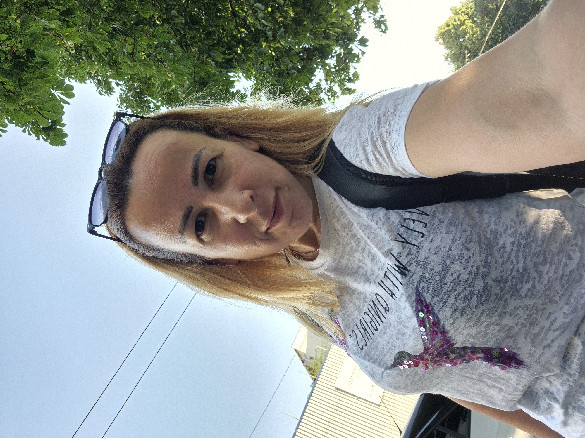 A woman with blonde hair and sunglasses perched on her head takes a selfie outdoors. She is wearing a light-colored t-shirt with a sequined hummingbird graphic and text that is partially obscured. She has a backpack on her shoulders. In the background, there is a building with a corrugated metal facade, some trees with green foliage, and power lines against a bright sky. The scene appears to be in an urban or suburban setting.