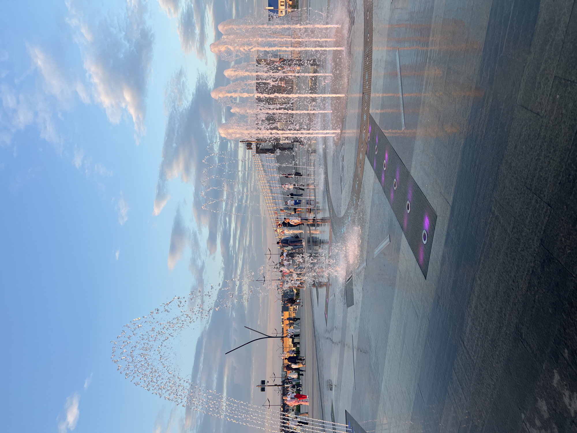 The image captures a wide shot of a public plaza with a synchronized fountain display at dusk in Magnitogorsk, Russia. Several people are visible walking and standing around the plaza, observing the water jets. The fountain features multiple streams of water shooting upwards, some illuminated with pinkish light, creating a dynamic visual effect. The plaza's surface is wet, reflecting the water and the ambient light. In the background, there are buildings and streetlights, indicative of an urban environment. The sky is a blend of blue and soft clouds, suggesting the transition from day to evening. The area appears to be a modern public space designed for leisure and recreation.