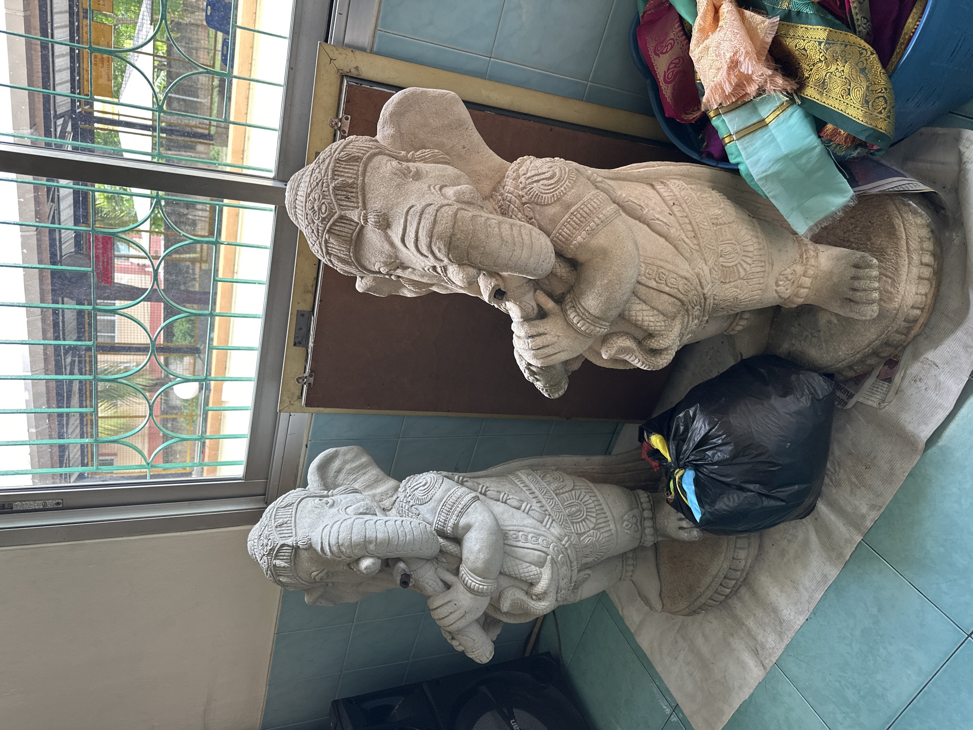 In the city of Unknown, Unknown, two stone statues of Ganesha, the Hindu deity, stand on a floor covered with a light-colored rug. The Ganesha statues are intricately carved, with one holding what appears to be a flute. To the left of the statues, a black trash bag is visible, and to the right, a blue bucket overflows with colorful textiles. Behind the statues, a large window with green bars offers a view of a courtyard with trees and buildings. The walls are painted a light color, with a blue tiled section below the window. A black speaker is positioned to the far left of the frame, and a framed object leans against the wall behind the statues.