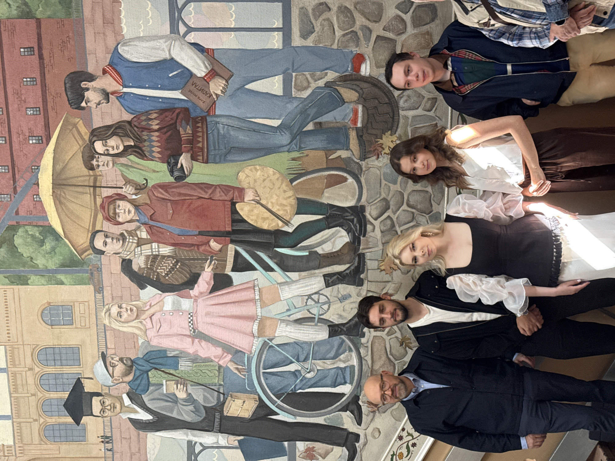 The image depicts a mural painted on a brick wall, possibly in the city of Saint Petersburg, Russia, given the style of architecture and the presence of what appears to be the Neva River and bridges in the background, common motifs in the city's art. The mural features a group of young people, rendered in a somewhat stylized, illustrative manner, standing on a cobblestone path. They are dressed in a mix of modern and retro-inspired clothing, suggesting a contemporary theme.
In the foreground, four individuals are standing in front of the mural. From left to right: a man with a shaved head and glasses, wearing a dark jacket over a blue shirt and dark trousers; a young man with dark hair, wearing a black jacket over a white t-shirt and black trousers; a young woman with blonde hair, wearing a black dress with puff sleeves and a white tiered skirt; and a young woman with long brown hair, wearing a white sleeveless top and brown trousers. To the right of the young woman in the white top is a young man with light brown hair, wearing a dark jacket over a striped shirt and tan trousers. To the far right, only the shoulder and arm of another person are visible, wearing a plaid shirt.
The mural itself shows a collection of figures, including one wearing a graduation cap, another holding a coffee cup, and a woman in a pink outfit with thigh-high socks and boots. A bicycle, an umbrella, and various accessories like a camera and a book are also depicted. The background of the mural shows buildings with arched windows and trees. The lighting in the foreground suggests an indoor setting, possibly a public space like a museum, gallery, or a communal area in a building.