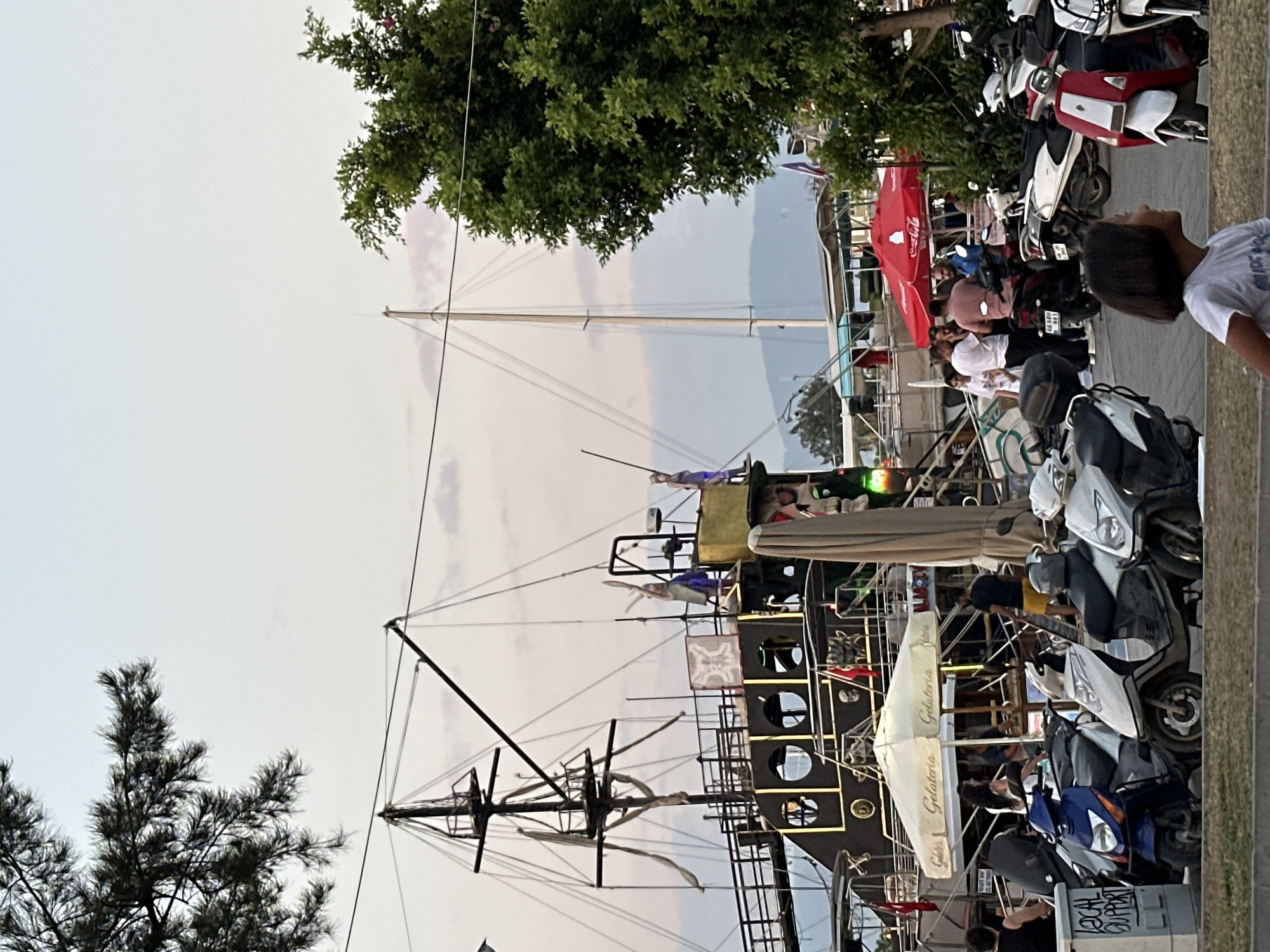 This is a medium shot, outdoor, eye-level photograph taken at dusk in Fethiye, Turkey. In the foreground, several parked scooters are visible, some of which are white, blue, and red. To the right of the scooters, a young girl with short, dark hair is looking to the right. Behind the scooters, a large, dark-colored boat with a pirate-ship aesthetic is docked. The boat has multiple decks, masts, and is adorned with yellow lights. A white umbrella with the text "Gelateria" is also visible near the boat. Several people are standing and walking around the boat and the area, including a man in a yellow shirt and black shorts, and a woman in a white top. In the background, a tall mast of another boat is visible, along with trees and mountains in the distance. The sky is overcast with faint pink hues, indicating sunset. A red Coca-Cola umbrella is also visible to the right.