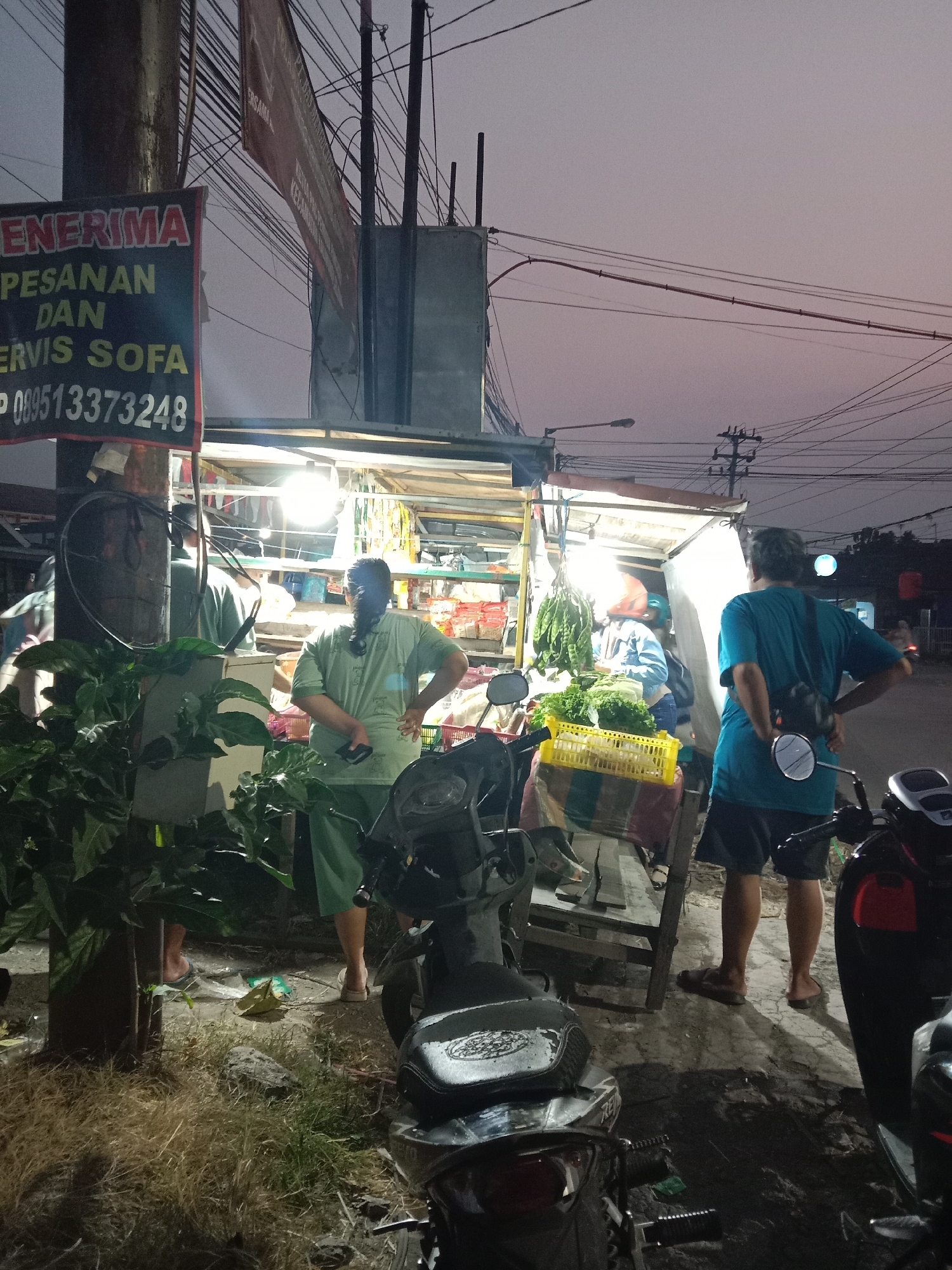 In Langensari, Indonesia, at dusk, a roadside vendor stall is illuminated by overhead lights. A banner reads "PENERIMA PESANAN DAN SERVIS SOFA" and displays a phone number. Three individuals are visible. Two men, one in a teal shirt and dark shorts, and another in a green shirt and pants, stand near the stall. The man in the teal shirt faces away from the camera, while the man in green faces left. A third person, whose gender is indeterminate, wearing a green shirt and pants, is seen from the back with a phone in hand. Behind them, the stall displays produce, including leafy greens and long, green vegetables. Motorbikes are parked in front of the stall, and the surrounding area features dry grass and power lines overhead.