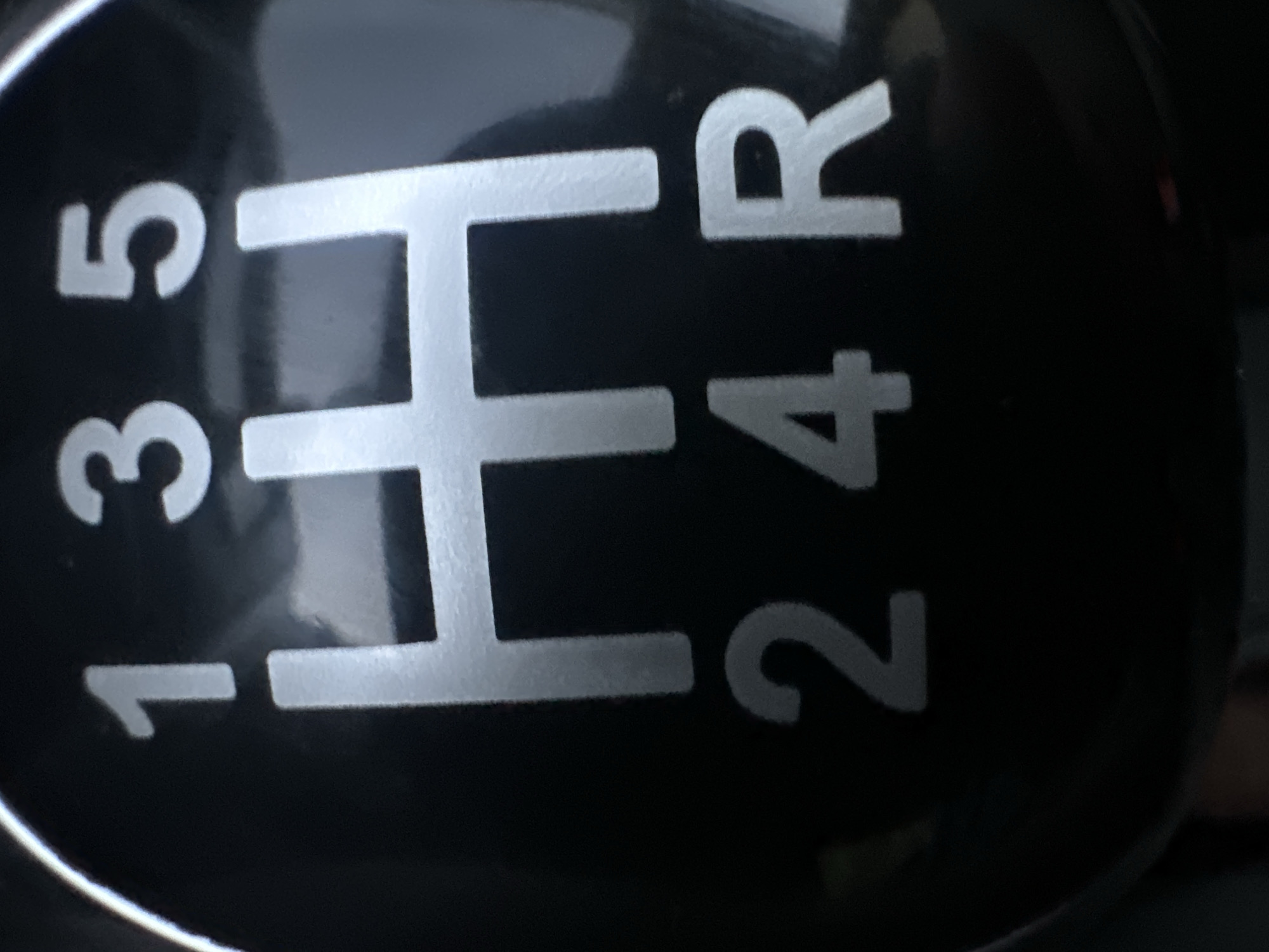 The image shows a close-up of a manual transmission gear shifter. The shifter is black with silver markings. The markings show a standard H-pattern for forward gears: 1, 3, and 5 are in the top row, while 2 and 4 are in the bottom row. The reverse gear (R) is also marked in the bottom row, to the right of the 4. The image is taken from a slightly elevated angle, looking down at the shifter. There are no people, actions, or interactions visible in the image. The environment is dark and appears to be the interior of a vehicle. The image does not contain any specific geographical markers or clues that would allow for its location to be determined in Plesetsk, Russia.