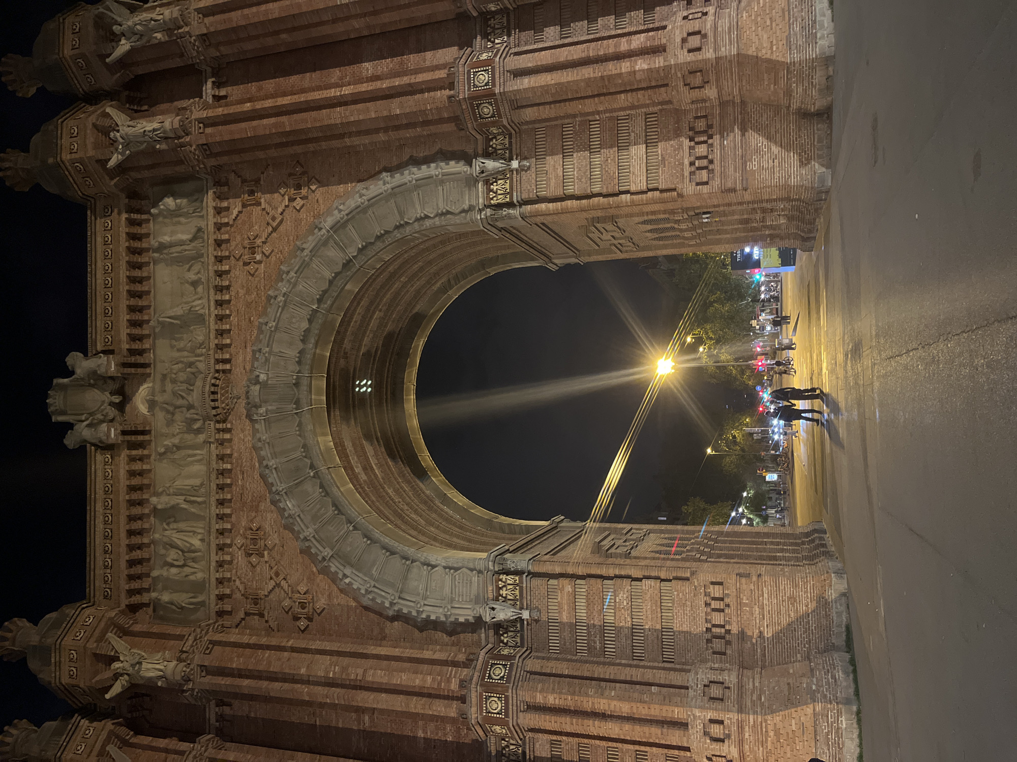 The image shows the Arc de Triomf in Barcelona, Spain, at night. The archway is made of red brick and is ornately decorated with sculptures and carvings. The archway frames a street with pedestrians walking under a streetlight.
