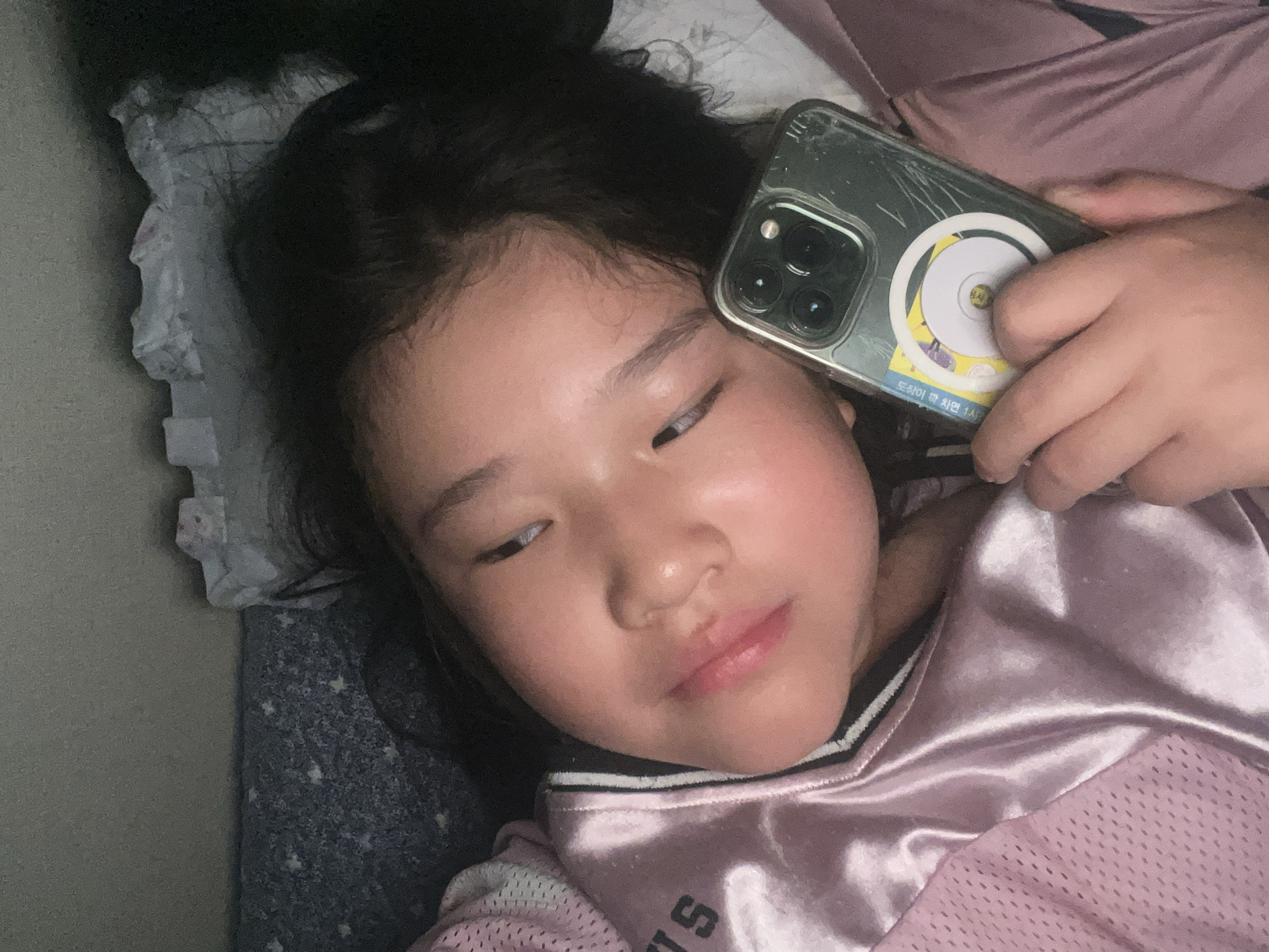 A young girl lies on a bed in Unknown, Unknown. She wears a pink jersey and rests her head on a pillow with a blue starry patterned sheet. She holds a cracked smartphone to her face, with a white circular attachment and a yellow sticker on the back.