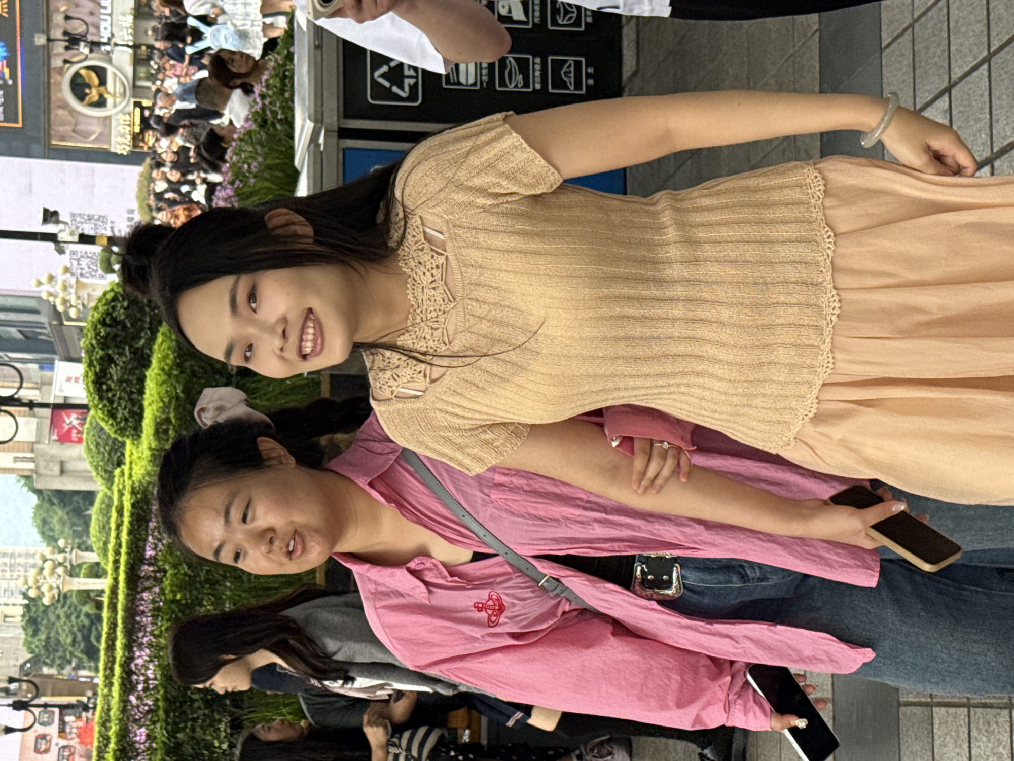 The image depicts two East Asian women in an urban setting, likely in Yuzhong District, China, based on contextual clues. The woman in the foreground wears a pink button-down shirt with a red Vivienne Westwood logo on it, blue denim jeans, and a gray shoulder bag. She is holding a black smartphone in her right hand. Behind her, the other woman is dressed in a tan knitted top with a lace neckline and a light peach-colored skirt. She is smiling slightly and appears to be aware of the camera. Both women have dark hair. In the background, there is a crowd of people, vegetation, and what appears to be a digital billboard with advertisements. There is also a recycling station. The setting suggests a bustling commercial area or pedestrian zone within the city.