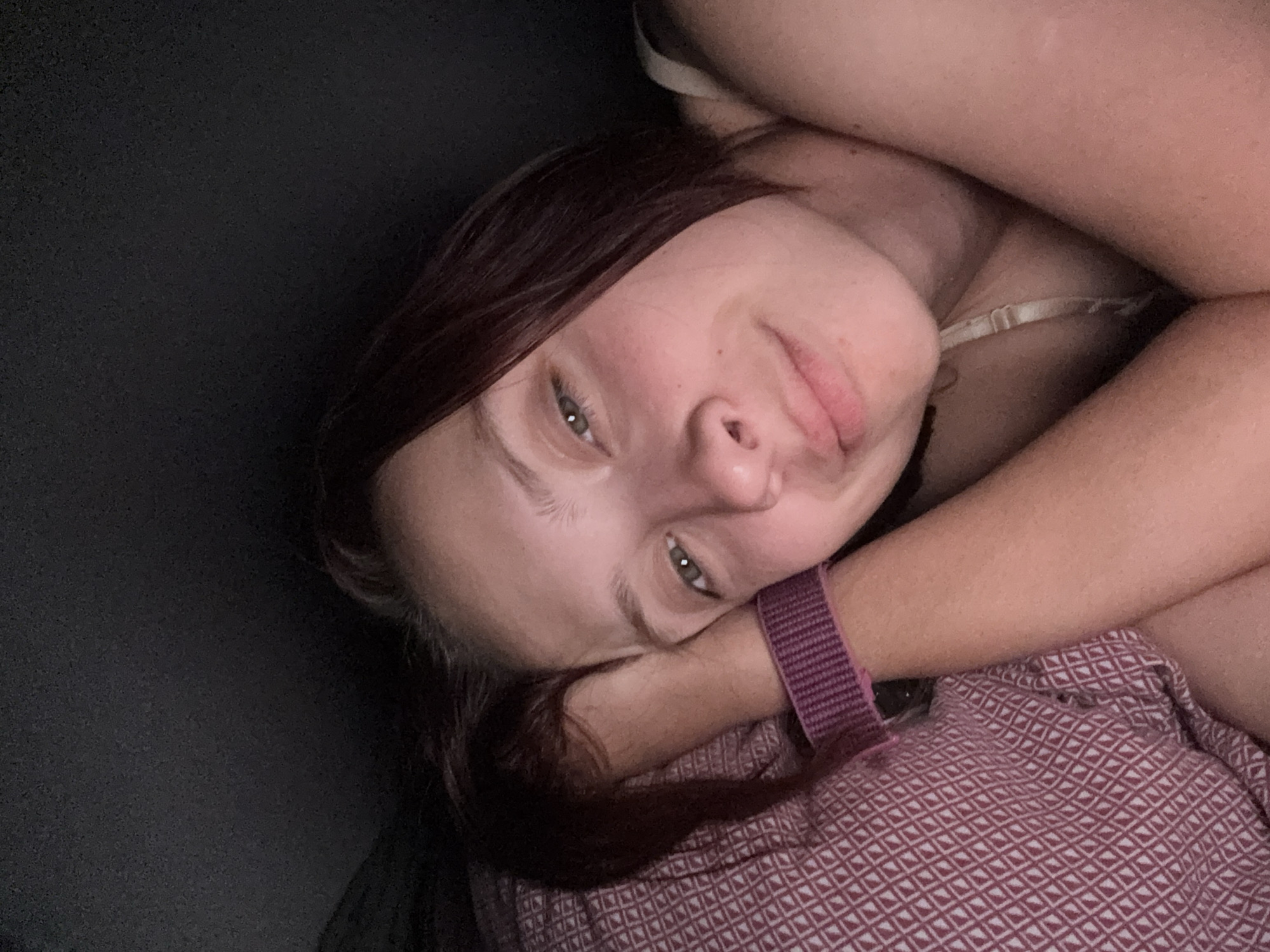 The image shows a close-up, selfie-style shot of a young woman lying down. She has auburn hair, fair skin with a smattering of freckles, and light green eyes. She is wearing a camisole with thin straps and a purple watch on her left wrist. Her head is resting on a pillow with a geometric pattern. The lighting is dim, suggesting it may be nighttime or indoors with low light. The background is dark and out of focus. There are no discernible landmarks or features that would indicate a specific location in Прикамский, Russia, or any city.