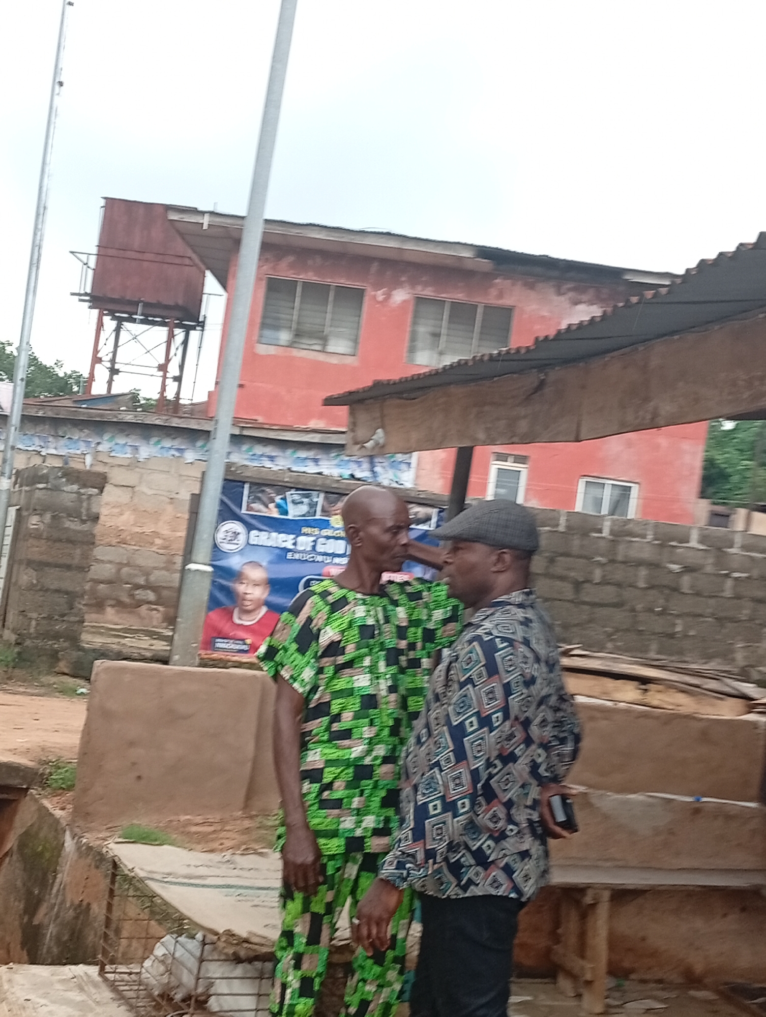 Two adult males are standing in an outdoor urban environment in Nsugbe, Nigeria. The male on the left is wearing a green and black patterned traditional African garment. He is facing towards the right and appears to be in mid-conversation with the other man. The male on the right is wearing a blue and white patterned shirt and a dark grey flat cap. He is facing towards the left, looking at the other man, and appears to be holding a black mobile phone in his right hand. Behind them, a red building with multiple windows is visible. To the left of the building, a red water tower on a metal frame stands tall. A blue banner with text and an image of a man is partially visible behind the men. The ground is covered with dirt and some debris. The sky is overcast.