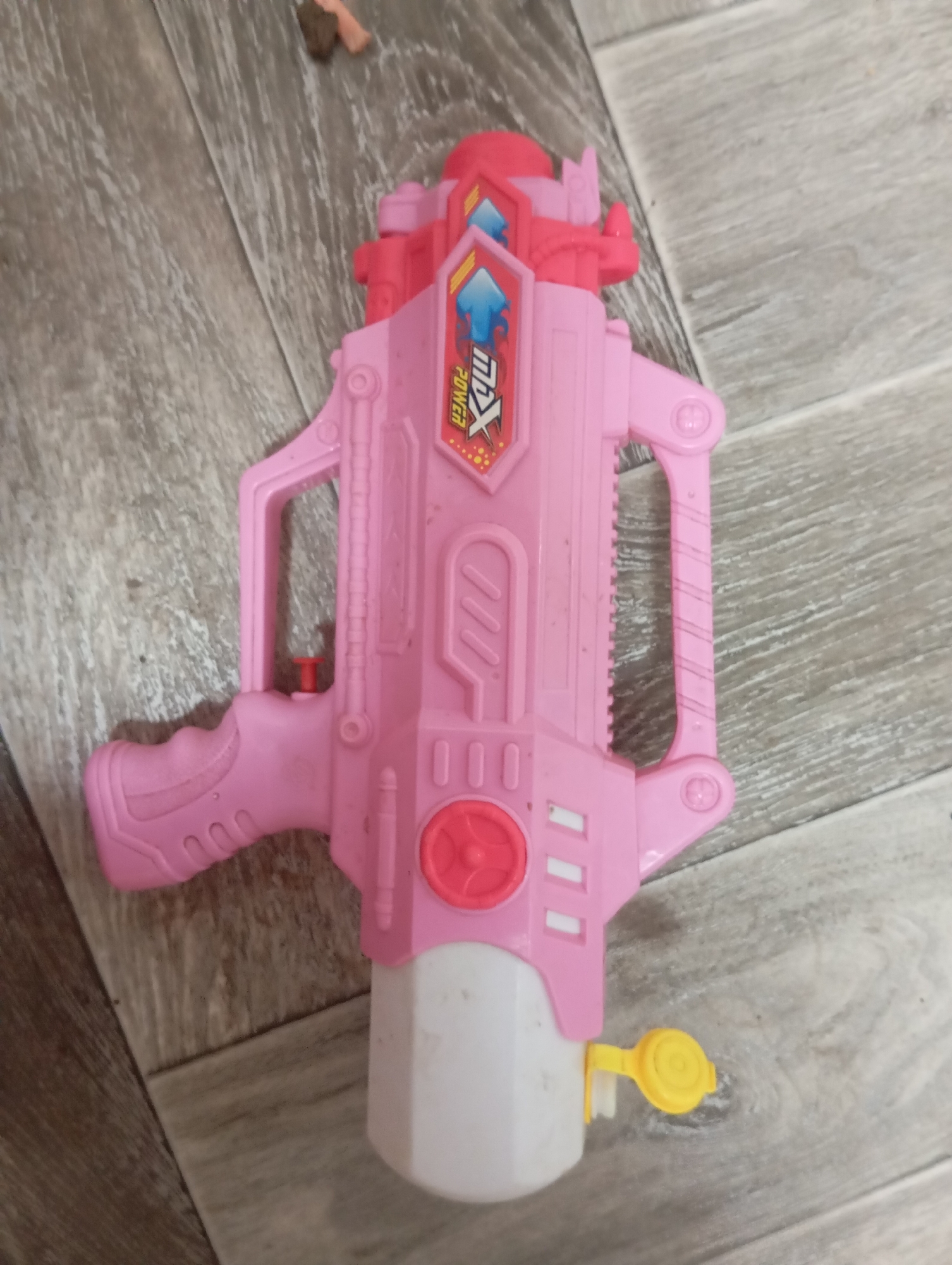 A pink toy water gun is lying on a wooden floor. The toy gun is made of plastic and has red accents. It has a handle, a trigger, and a barrel. There is a white reservoir at the bottom of the gun with a yellow cap. The gun is decorated with a shield-shaped emblem that reads "MX POWER" in red and blue letters. The wooden floor is made of planks of varying shades of brown and gray. The scene appears to be indoors.