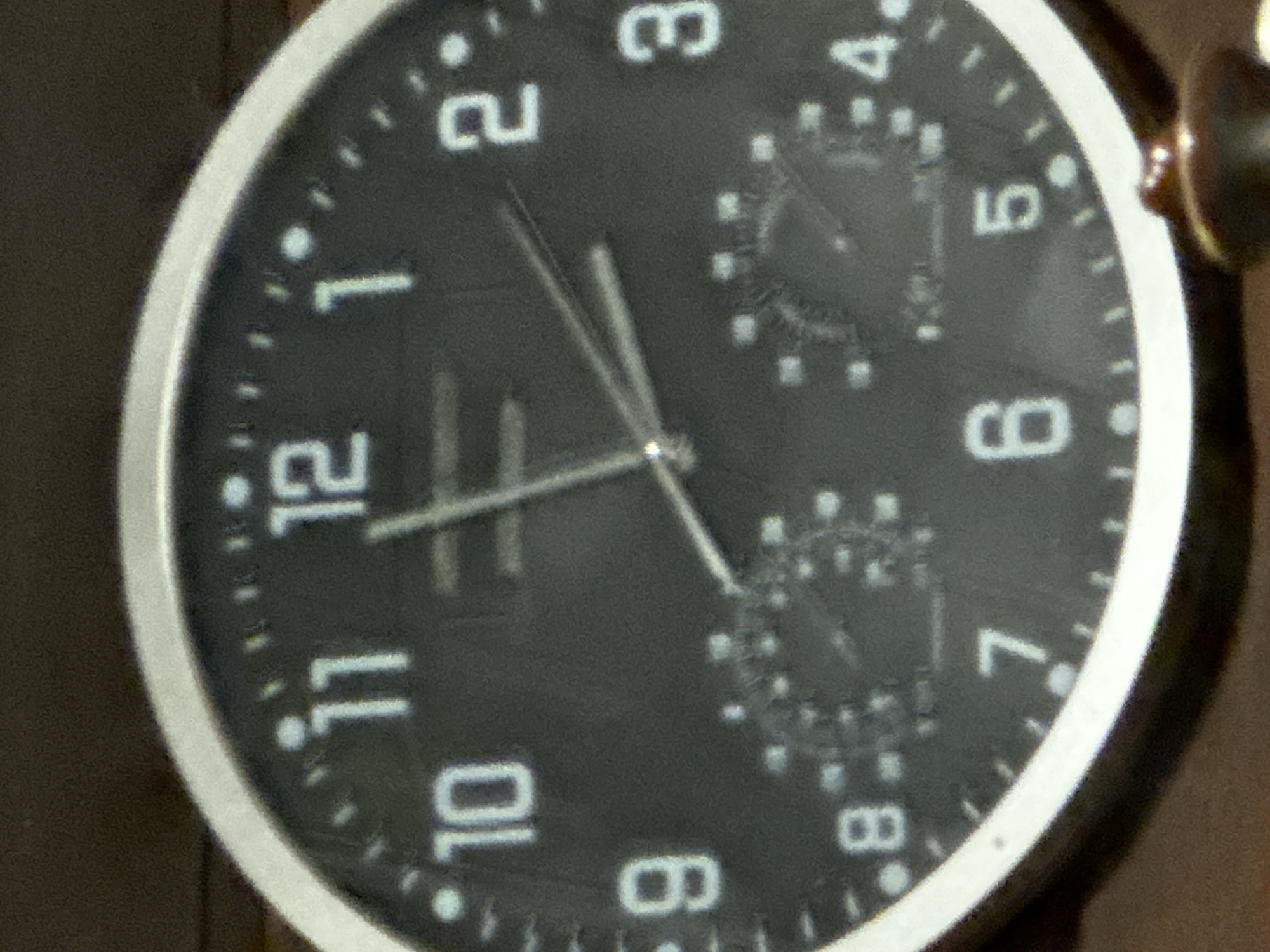 This is a close-up, slightly blurry, medium-shot photograph of a black analog clock face with white numerals. The clock has a silver bezel. The numbers on the clock face are 1 through 12. The hour hand is pointing between the 11 and the 12, and the minute hand is pointing just past the 12. There are also two smaller dials on the clock face, located around the 7 and the 5 positions, which appear to be sub-dials for a chronograph or other functions, though their readings are unclear due to the image quality. The clock is mounted on a brown wooden surface, and there is a dark, out-of-focus background. The specific location in "Востец Снт, Russia" is not discernible from the image itself.