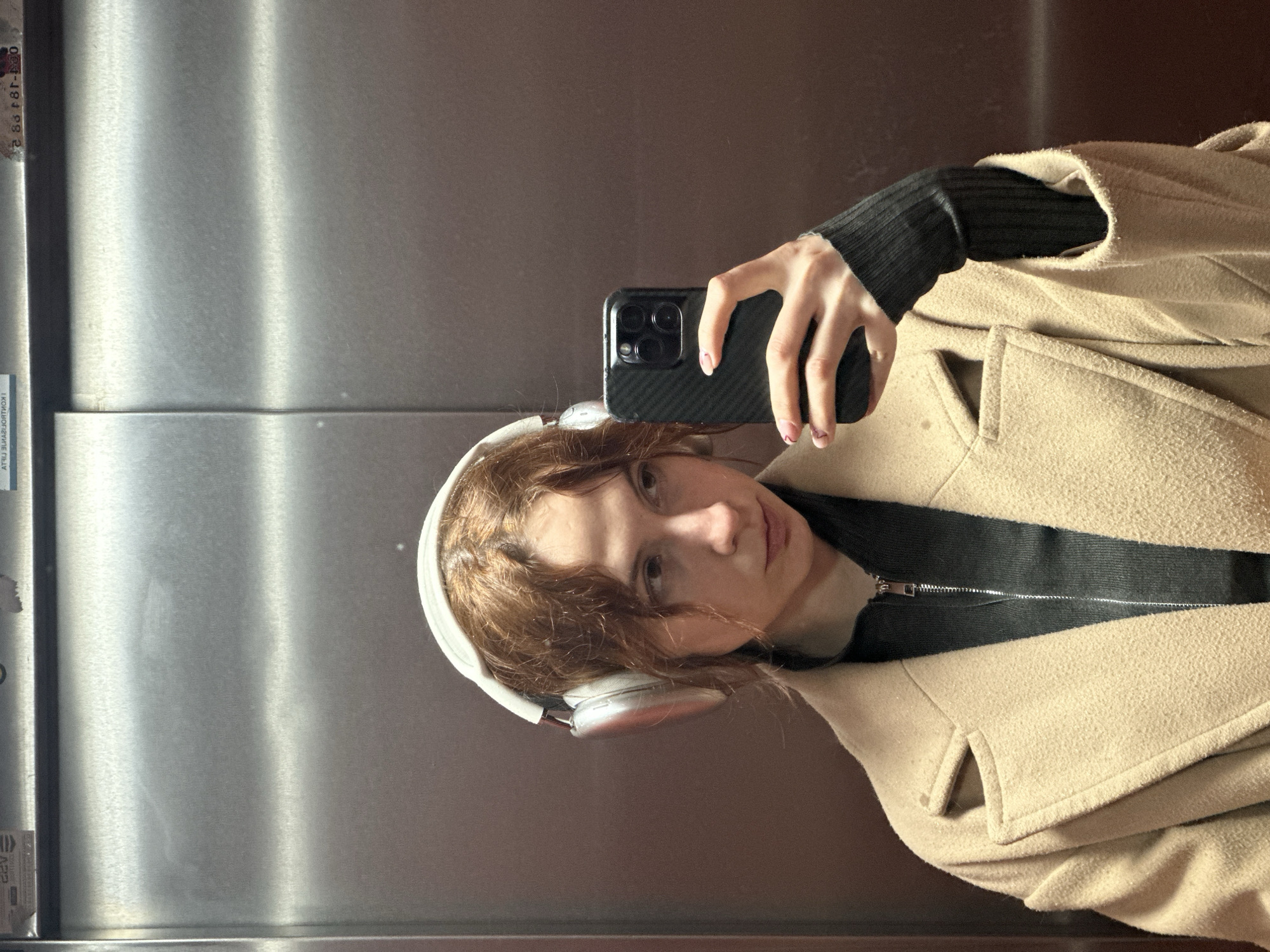 The image shows a person taking a selfie in an elevator with a metal interior. The person has fair skin, brown curly hair, and is wearing white headphones. They are wearing a black long-sleeved shirt or sweater underneath a beige coat. The person is holding a black smartphone with a multi-lens camera system. Based on the interior of the elevator, the image was taken in Novi Sad, Serbia.