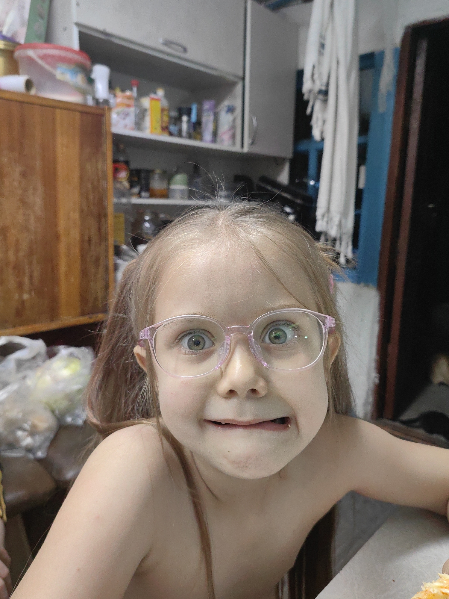 This is a close-up, eye-level, medium-shot photo of a young Caucasian girl with blonde hair in pigtails, wearing pink-rimmed glasses. She is looking directly at the camera with a playful, slightly cross-eyed expression and her mouth is pursed, showing her teeth in a funny grimace. The girl is shirtless.
The background is blurred but appears to be a domestic setting, possibly a kitchen or pantry. To the left, there is a wooden cabinet or cupboard, and above it, shelves are visible containing various jars and containers. To the right, there is a doorway with a dark interior and a blue vertical element, possibly part of a door or cabinet. White curtains or fabric hang behind the girl on the right side. There are also plastic bags with some items inside visible on a table or counter to the left and in the lower portion of the image.
The lighting suggests it is daytime, but the indoor setting makes it difficult to determine the exact time or weather. The overall mood conveyed by the girl's expression is one of fun and playfulness. No text is visible in the image.
