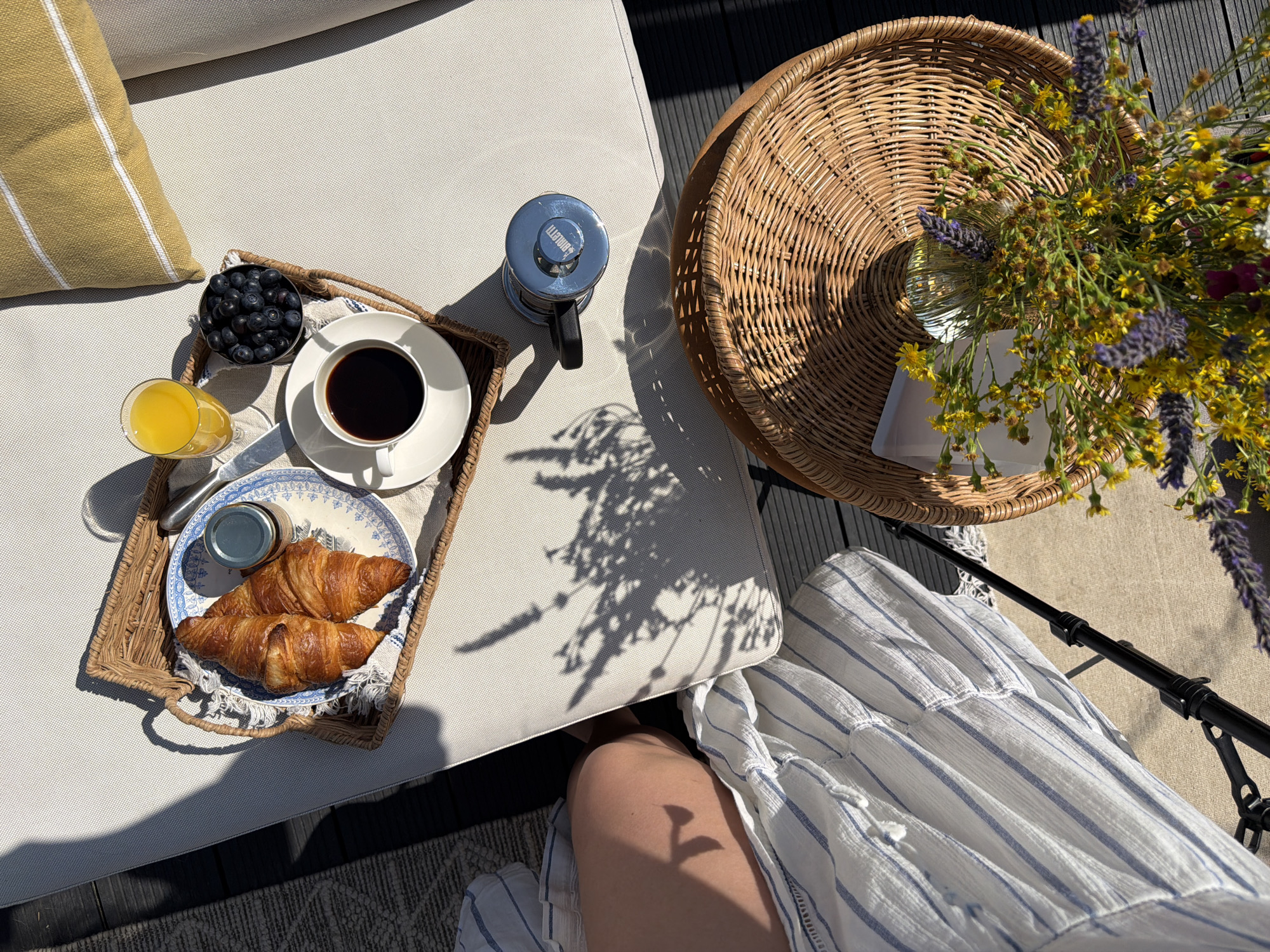 This image depicts a close-up, overhead view of a breakfast spread arranged on a white outdoor lounge chair. The scene is set in Gangloff, Germany.
In the foreground, a woven basket contains a bouquet of wildflowers, predominantly yellow with hints of purple and green. A white piece of paper is visible peeking out from under the flowers.
To the upper left, a wicker tray holds a breakfast for one. Two golden-brown croissants are placed on a blue and white patterned plate, next to a small jar with a silver lid. A white teacup filled with dark coffee sits on a saucer, to the right of the croissants. A glass of orange juice is positioned above the plate, and a small metal bowl filled with blueberries is to the right of the coffee cup. A chrome French press coffee maker is situated on the white surface of the lounge chair, to the right of the tray.
A person, wearing a white dress with thin blue stripes, is partially visible on the far left of the image, with their legs crossed. The lounge chair is positioned on a dark wooden deck. The scene is bathed in bright sunlight, casting distinct shadows from the flowers and the tray onto the chair and the deck. A yellow pillow with white stripes is visible at the top right corner of the image.