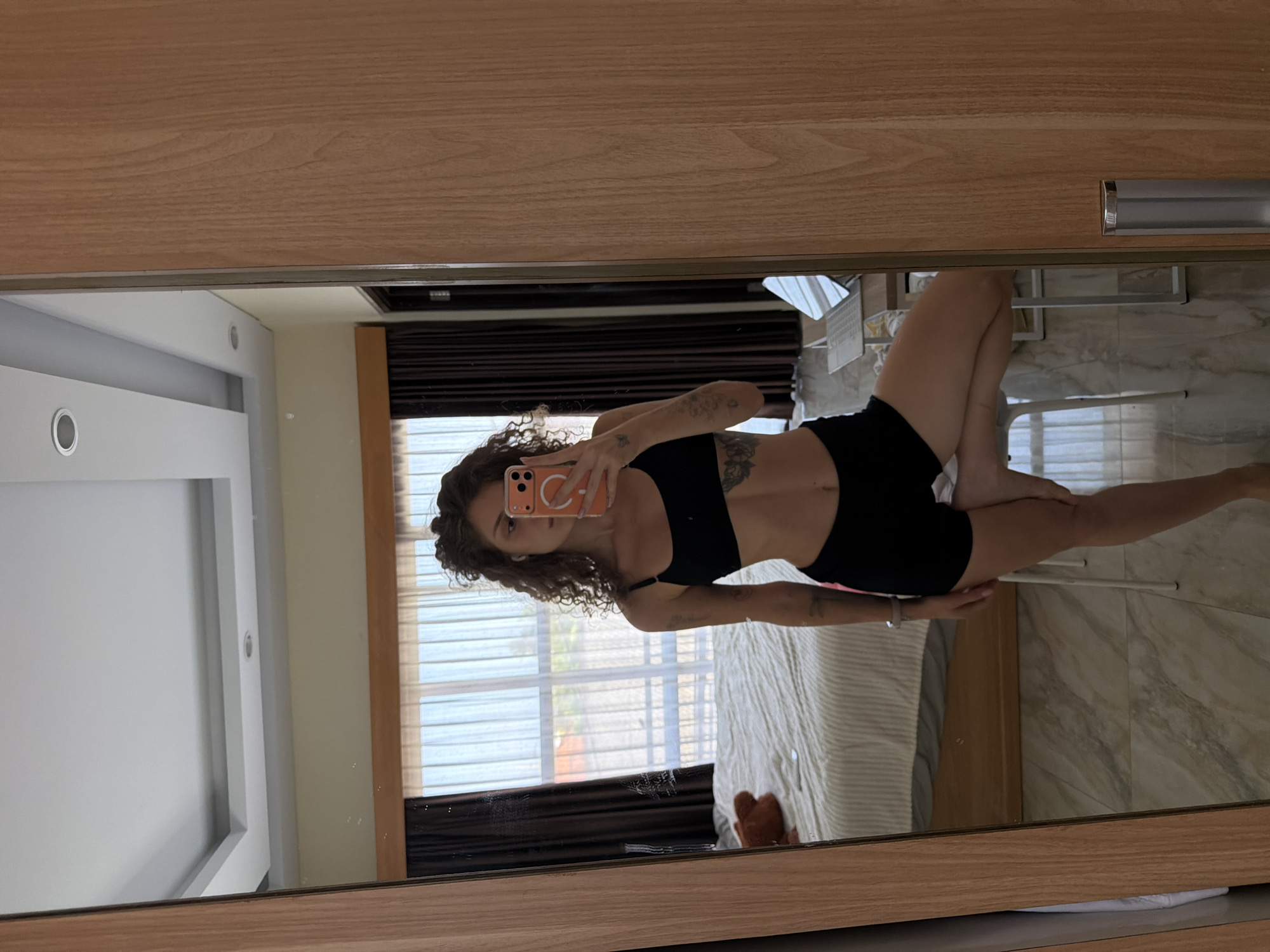 A young woman with curly brown hair is taking a mirror selfie in a bedroom in North Kuta, Indonesia. She is wearing a black sports bra and black shorts. She is holding up an orange smartphone to take the picture. In the background, a bed with a white and beige striped comforter is visible. There are dark brown curtains on the windows, and sheer white curtains hang in front of them. A laptop is on a small table to the right of the bed. The room has light-colored tile flooring and a white ceiling with recessed lighting.