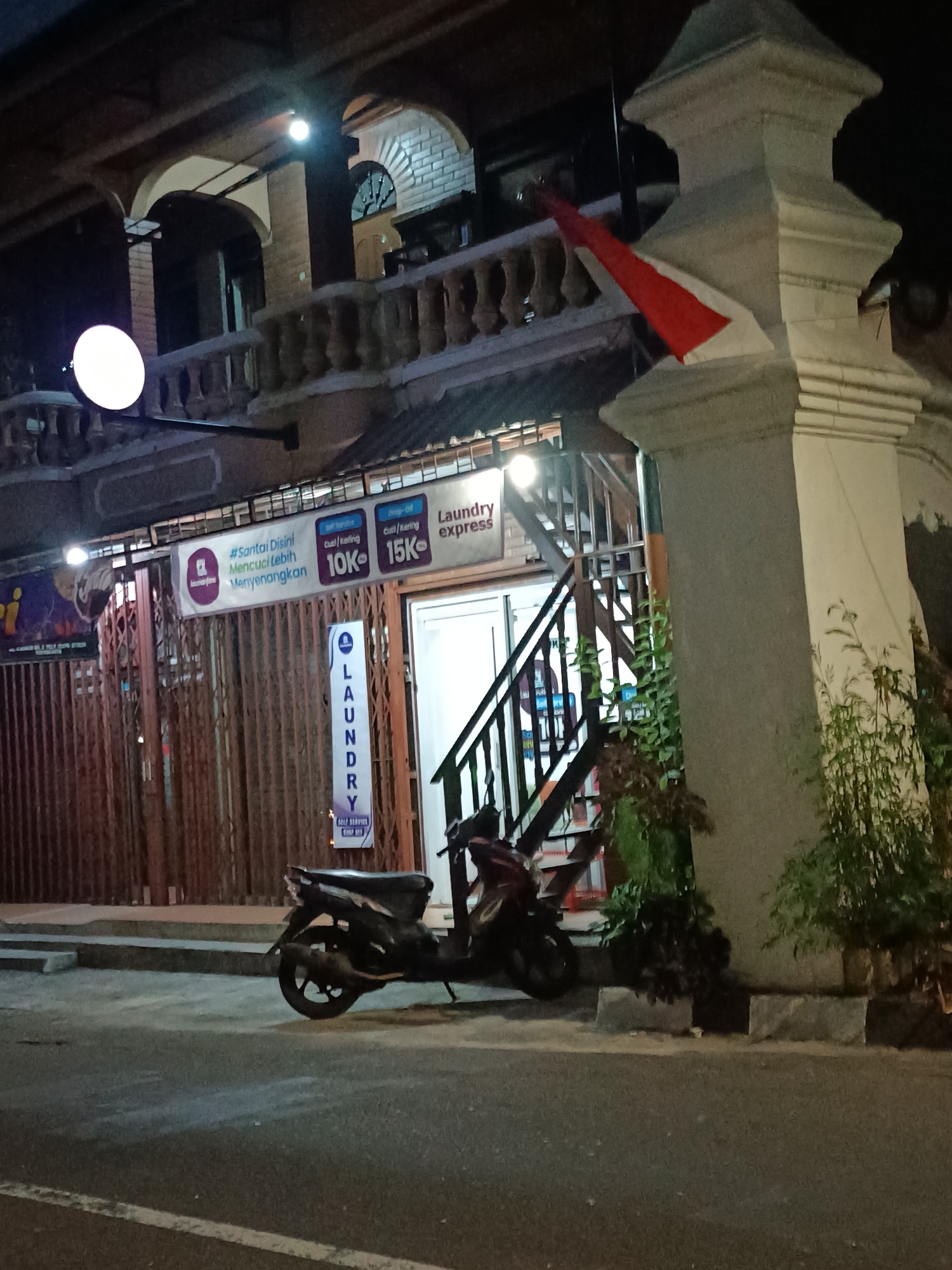 The image shows a laundry shop with an Indonesian flag draped over a pillar. The shop is located in Yogyakarta, Indonesia. The shop has a modern design with a glass storefront and a metal grille. There are two motorcycles parked in front of the shop. The street is lit by streetlights, and the shop is illuminated by neon signs. The image is taken at night, and the sky is dark.