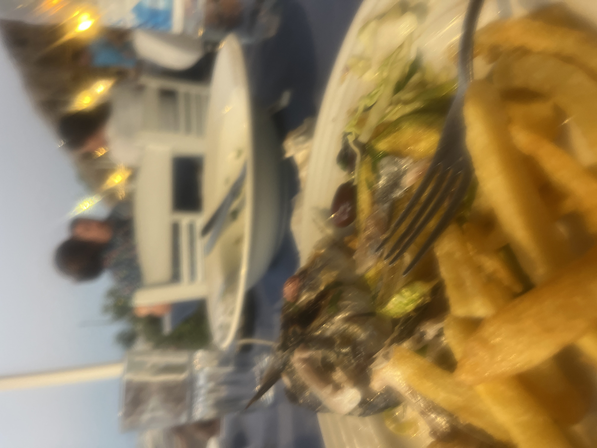 An out-of-focus, close-up view of a plate of food and a table setting at what appears to be an outdoor restaurant in Polis Chrysochous, Cyprus. The plate contains a partially eaten fish, french fries, and a salad with green leafy vegetables and sliced zucchini. A fork rests on the food. In the background, blurred figures of people are visible, along with tables and chairs. The lighting suggests it is either late afternoon or early evening, with some artificial lights also present. The overall impression is of a casual dining experience.