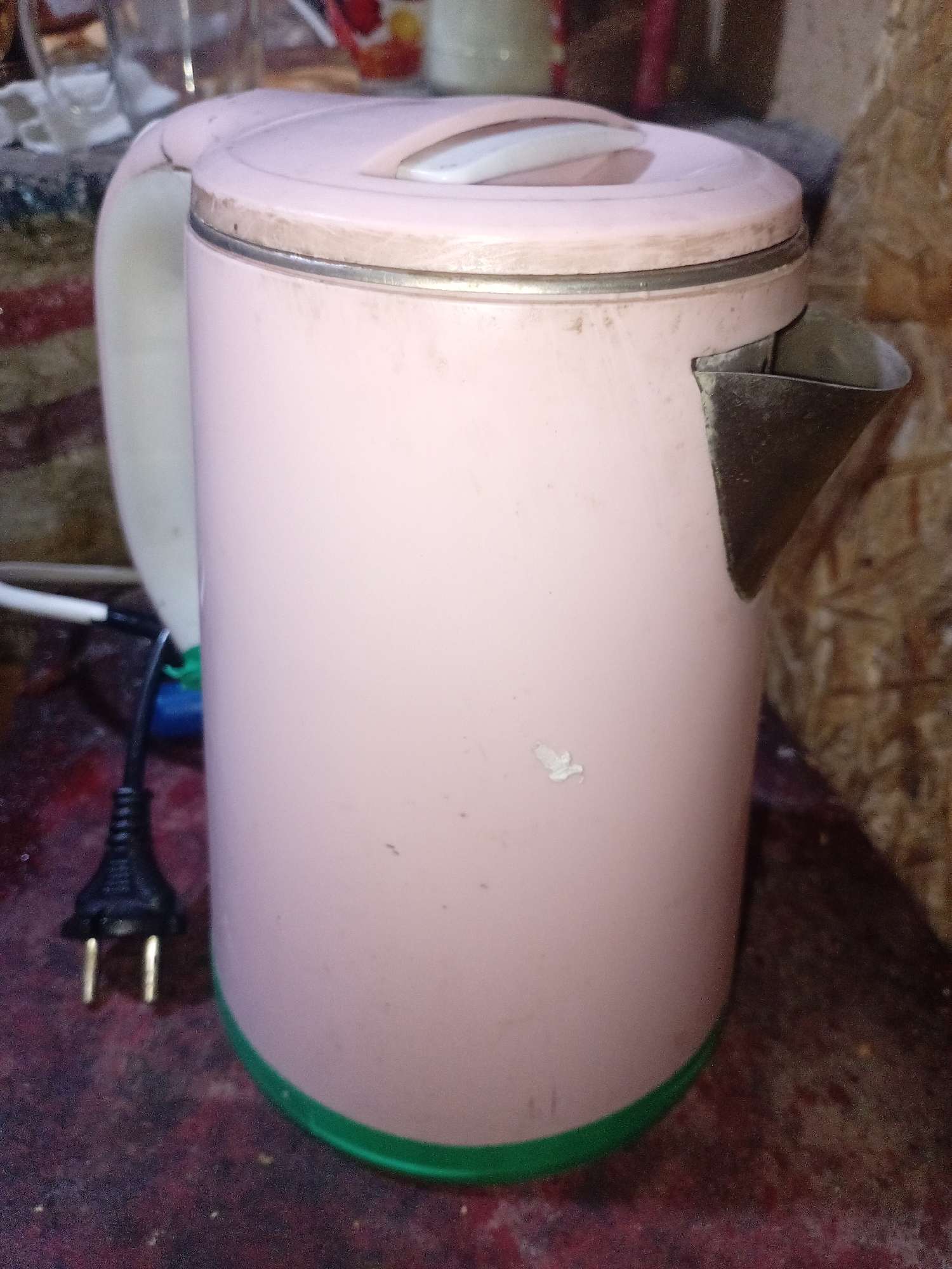 A pink electric kettle with a green base and a metal spout sits on a textured, dark red surface. A black electrical plug with two prongs is visible to the left of the kettle. The background appears to be a wooden wall. The scene is set in a cluttered environment, possibly a kitchen or workshop, in Zeravshan-2, Tajikistan.