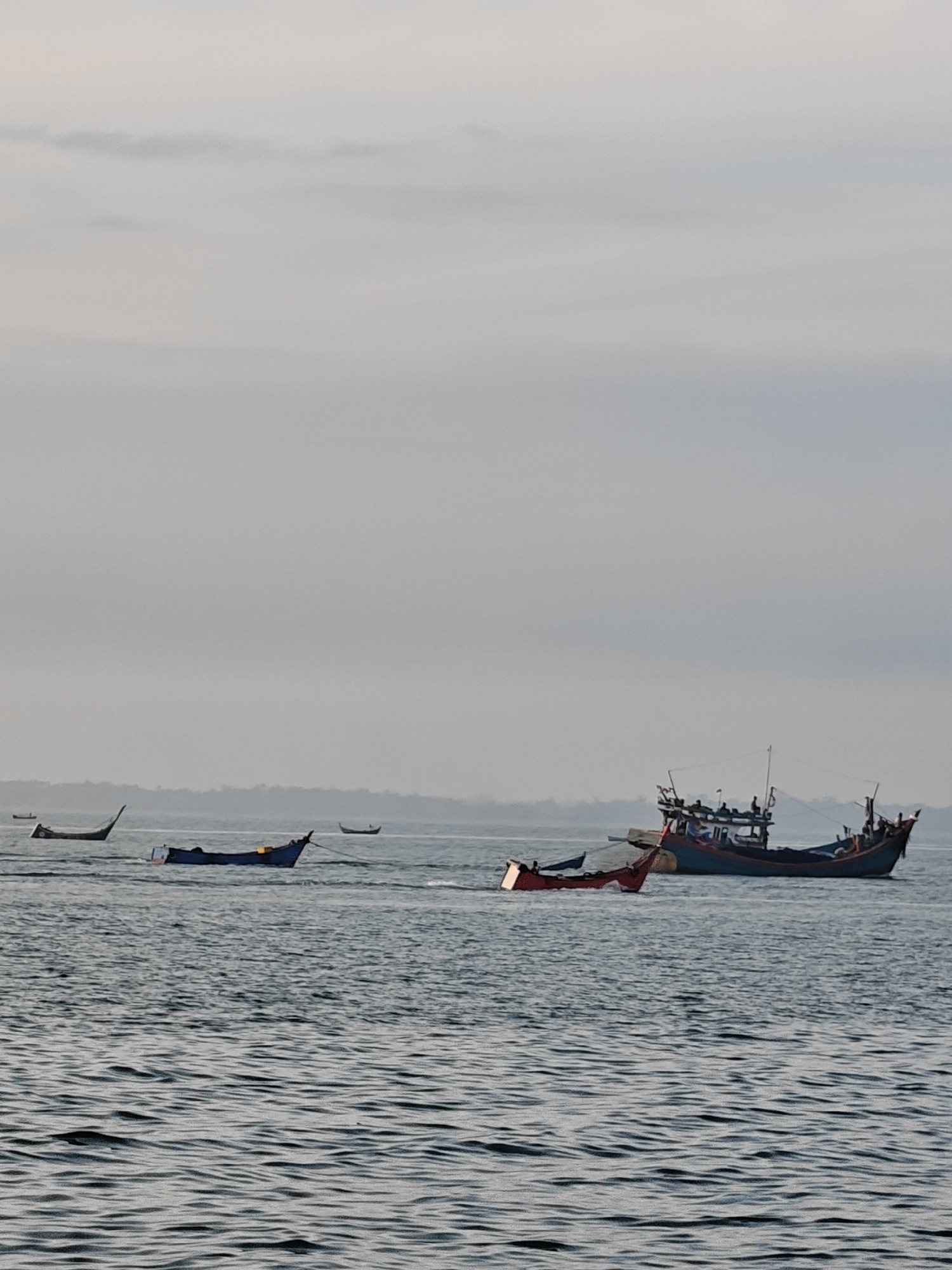 The image shows a wide shot of the ocean near Lhokseumawe, Indonesia. A large fishing boat with a blue and red hull is the central subject, positioned on the right side of the frame. The boat has a multi-level structure with a cabin and appears to be carrying several people. In front of the large boat, a smaller red boat is visible, cutting through the water. To the left of the large boat, a blue fishing boat is anchored. Further to the left, two more small boats are seen in the distance, one with a curved hull and another smaller dark boat. The sea is choppy with visible waves. The sky is overcast and light gray. In the background, a hazy, tree-lined coastline is visible, suggesting the proximity of land. The overall atmosphere is serene and depicts a typical day at sea for fishermen.
