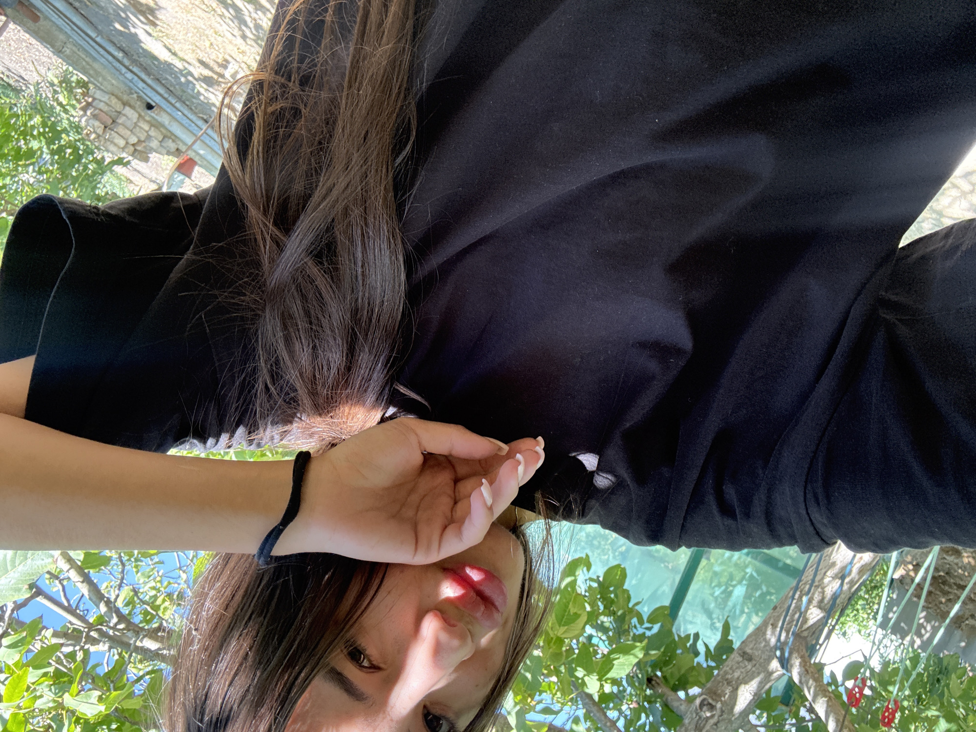 A woman with dark hair is taking a selfie outdoors, leaning her head on her hand. She is wearing a black t-shirt and a thin black bracelet on her wrist. Her lips are slightly parted and painted red. In the background, there is a tree with green leaves, and a hazy view of what appears to be a greenhouse or protective netting. The overall setting suggests an outdoor area, possibly a garden or yard. The exact location in Basarabeasca, Moldova, is not discernible from the image itself.
