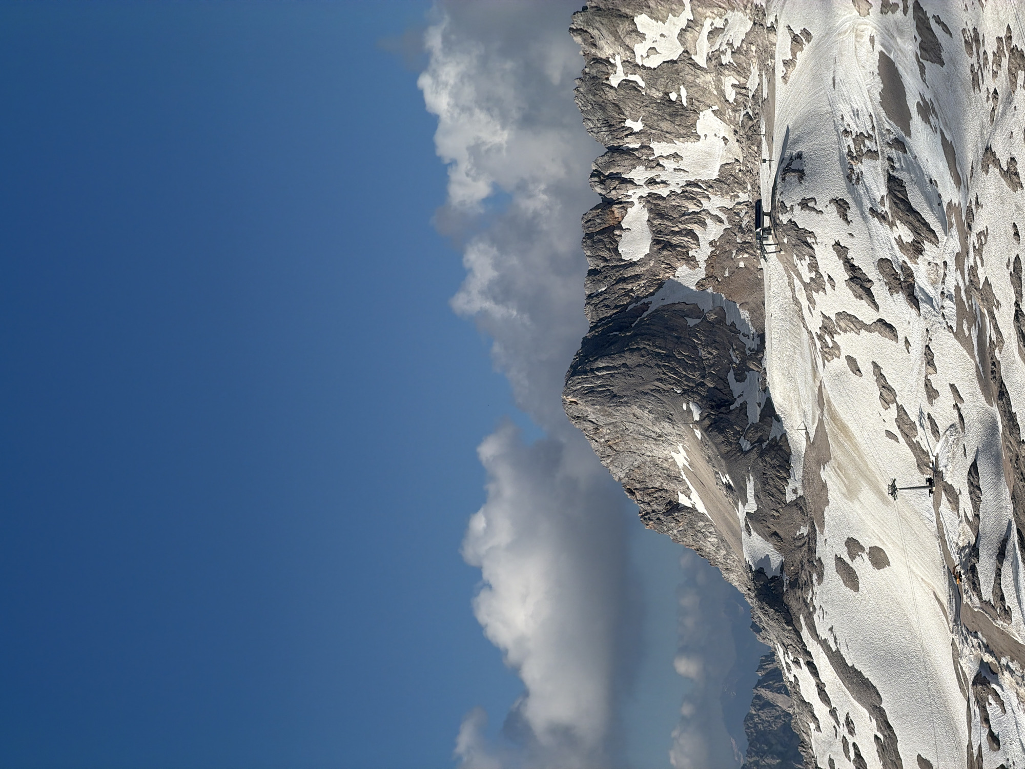 The image shows a high-altitude mountain landscape with a bright blue sky and scattered clouds. The mountains are rugged and rocky, with patches of snow covering the slopes. A cable car system is visible, with a car suspended from a cable traversing the snowy mountainside. A yellow construction vehicle is also present on the snowy terrain. The lighting suggests it is daytime, likely mid-morning or afternoon, with sunlight casting shadows across the landscape. The scene evokes a sense of remote wilderness and outdoor adventure. No visible text is present. The location context provided is Garmisch-Partenkirchen, Germany, which is known for its mountainous terrain and ski resorts.