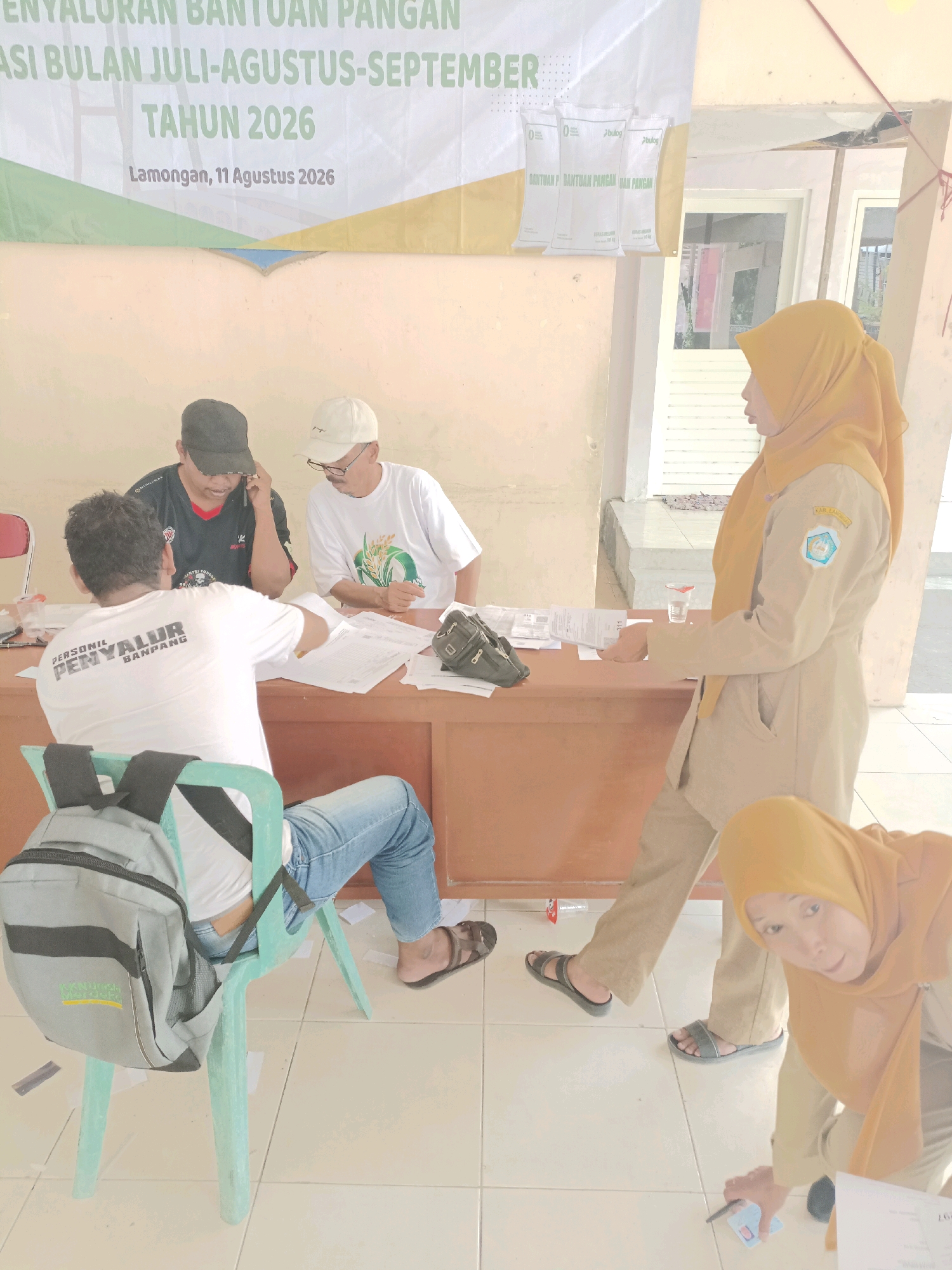 The image depicts an indoor scene, likely a community hall or office, in Guminingrejo, Indonesia. A banner in the background announces "PENYALURAN BANTUAN PANGAN" (Food Aid Distribution) for July-August-September 2026, held in Lamongan on August 11, 2026.
There are four individuals visible. Two men are seated at a wooden desk, engaged with papers. One man, wearing a dark baseball cap and a red and black t-shirt, is on the phone. The other, wearing a white cap and glasses, is looking at the papers. A third man, from the back, is seated on a light green plastic chair, wearing a white t-shirt with text "PERSONIL PENYALUR BAN Pang" and blue jeans, with a gray backpack on the chair. A woman in a tan uniform with a yellow headscarf is standing, facing the desk, and appears to be interacting with the documents. Another woman, also in a tan uniform and yellow headscarf, is partially visible in the foreground, kneeling and looking at the camera, holding a pen and a card.
The environment appears functional, with the wooden desk and papers suggesting an administrative process. The tiled floor is visible, and some papers are scattered on it. The overall lighting is soft, indicating an indoor setting with ambient light.