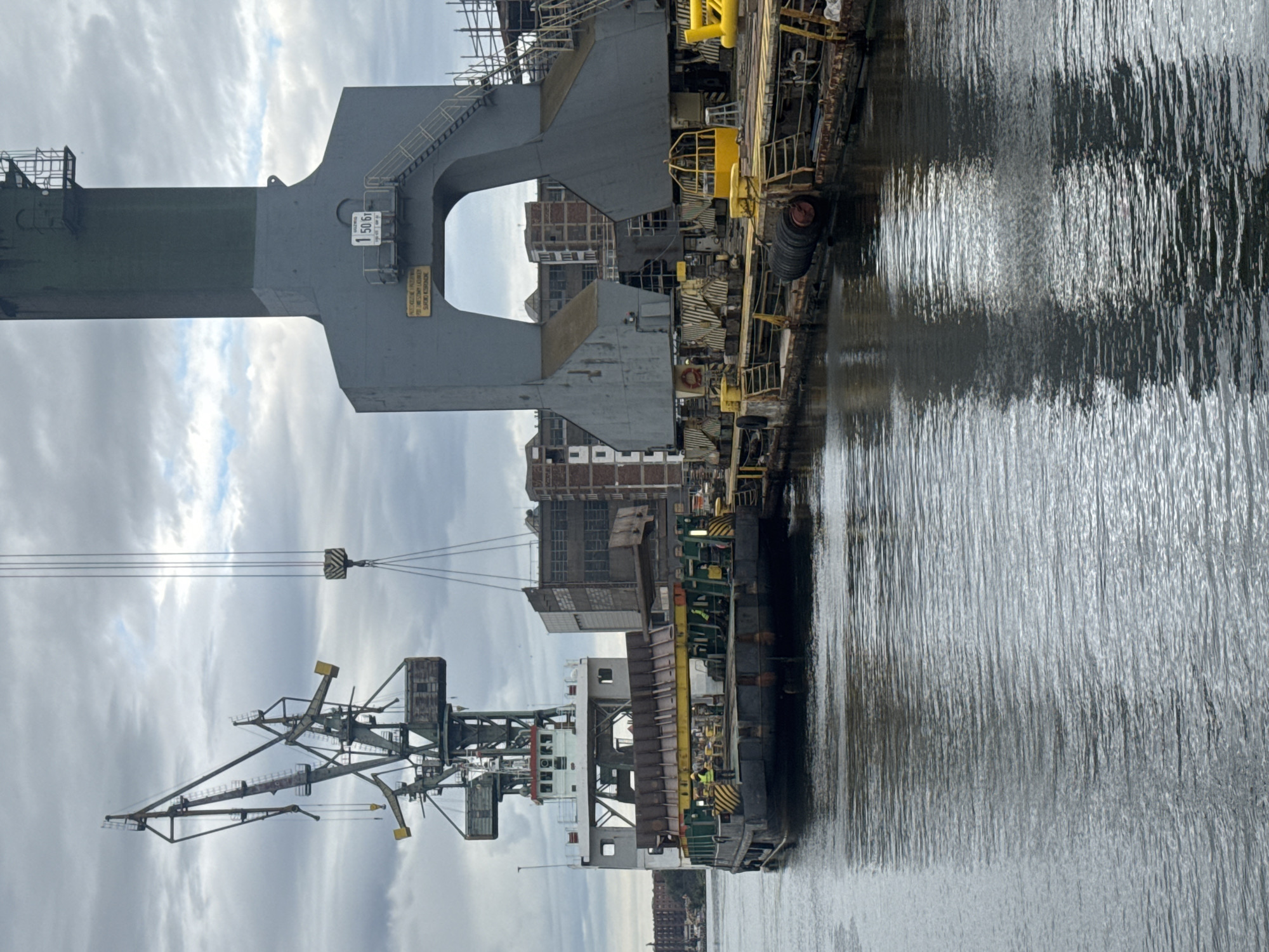 The image depicts an industrial scene at the Gdańsk Shipyard in Gdańsk, Poland. A barge is docked next to a large grey crane structure, with a smaller crane visible in the background. The barge, loaded with metal components, is positioned to receive or offload material. Several individuals, some wearing high-visibility vests, are present on the barge and the dock, suggesting ongoing work. The environment is a waterfront with calm water reflecting the overcast sky and industrial structures. The large crane has a sign indicating a weight capacity of "1 50 6t" and warning text in Polish. The scene suggests heavy industrial operations within the shipyard.