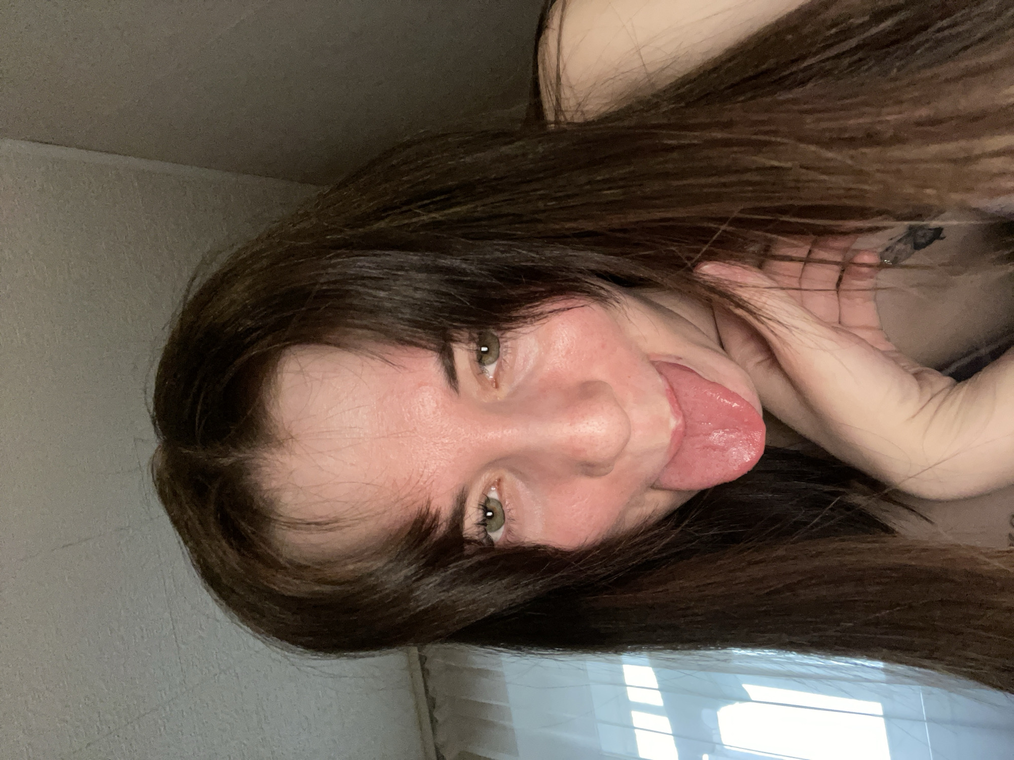 A woman with long, dark hair is sticking her tongue out towards the camera. Her eyes are green. She is indoors. It is not possible to determine the location as Chaikovsky, Russia based on the image.