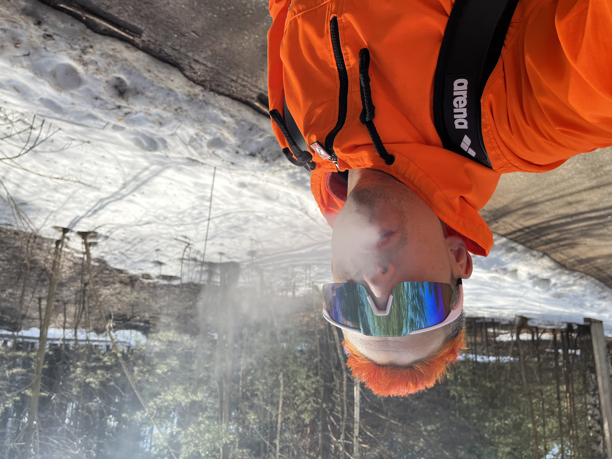 A man is shown outdoors in an upside-down, medium shot. He has bright orange hair and is wearing white-framed mirrored sunglasses and an orange jacket, on which the word "arena" is visible. He is exhaling, and the condensation from his breath is visible. The background suggests a wooded area with a layer of melting snow on the ground. The image was taken in Chaikovsky, Russia.